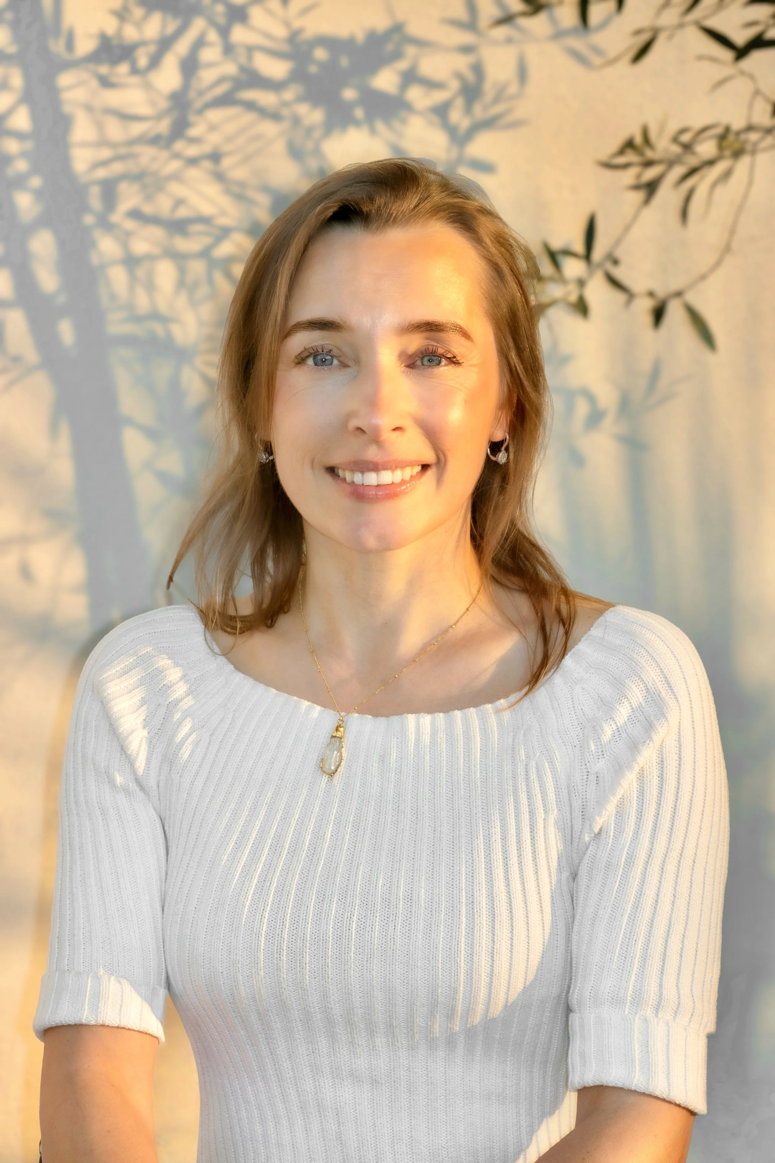 A woman with shoulder-length light brown hair and blue eyes smiling, wearing a white ribbed sweater and jewelry, with shadows of tree branches and leaves on a wall behind her.