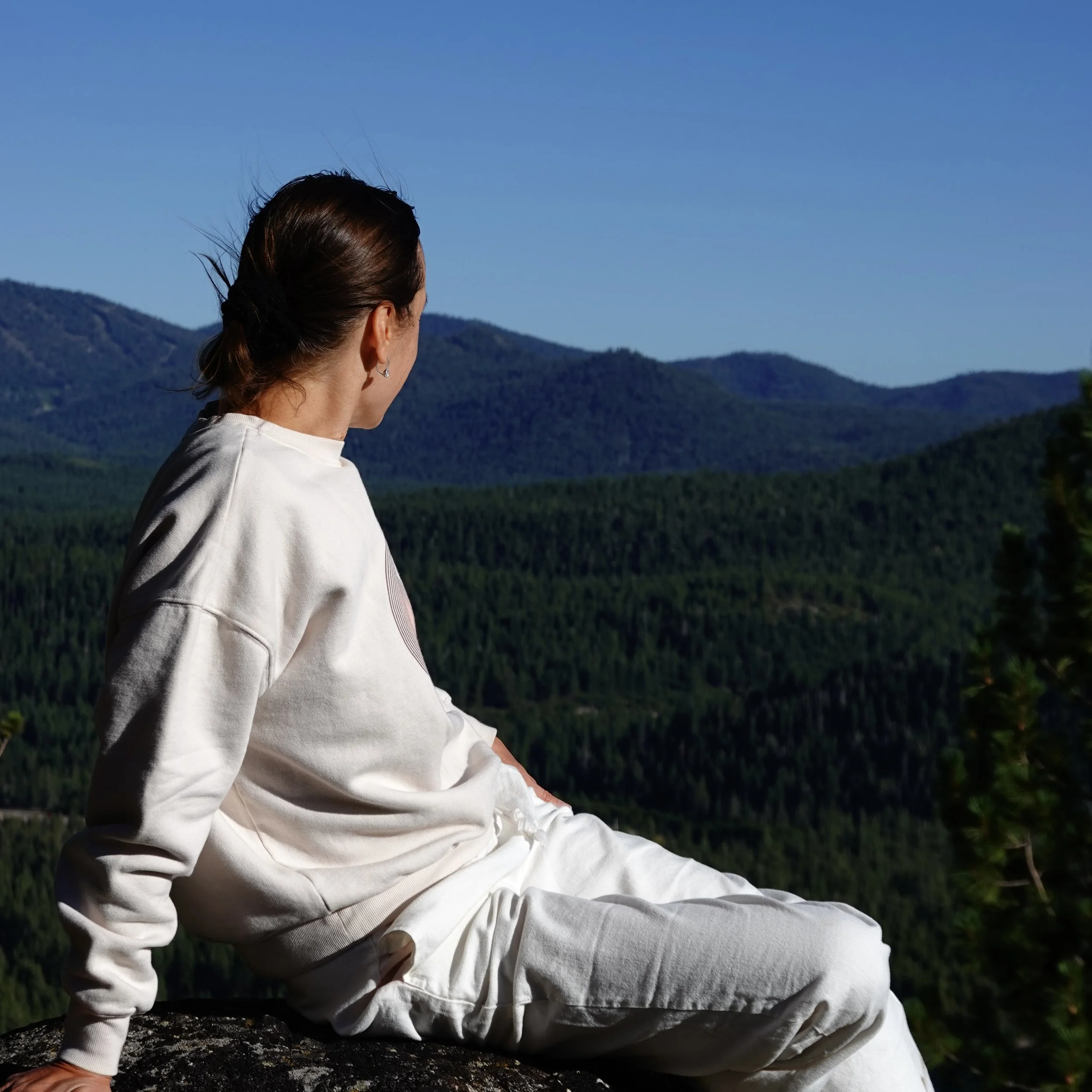 A woman with brown hair tied back, wearing a white sweatshirt and white pants, sitting outdoors on a rock with a scenic view of green mountains and clear blue sky in the background.