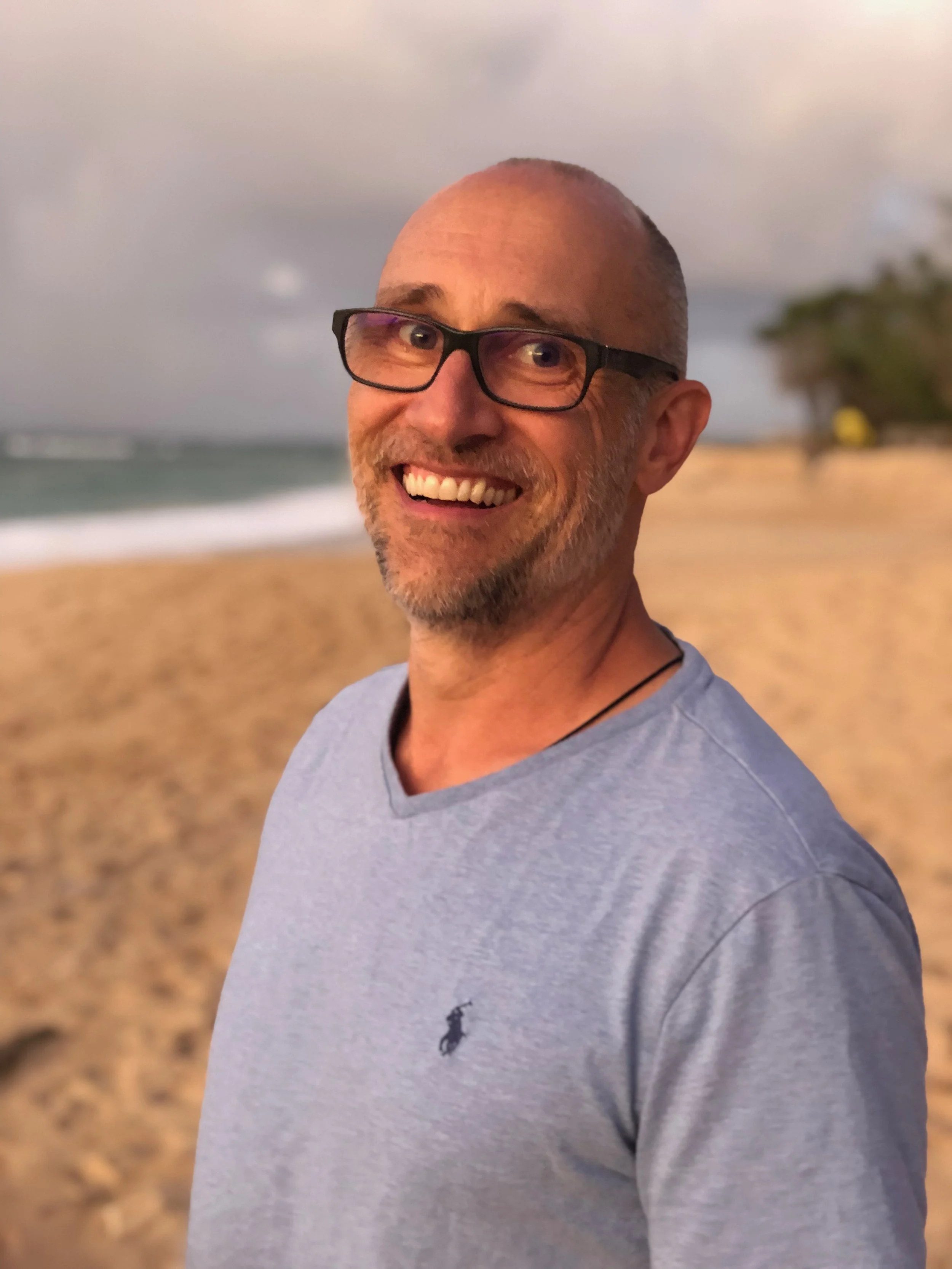 A smiling man with glasses and a short beard outdoors on a beach. In the background, there are blurred trees, sand, and the ocean under a cloudy sky.