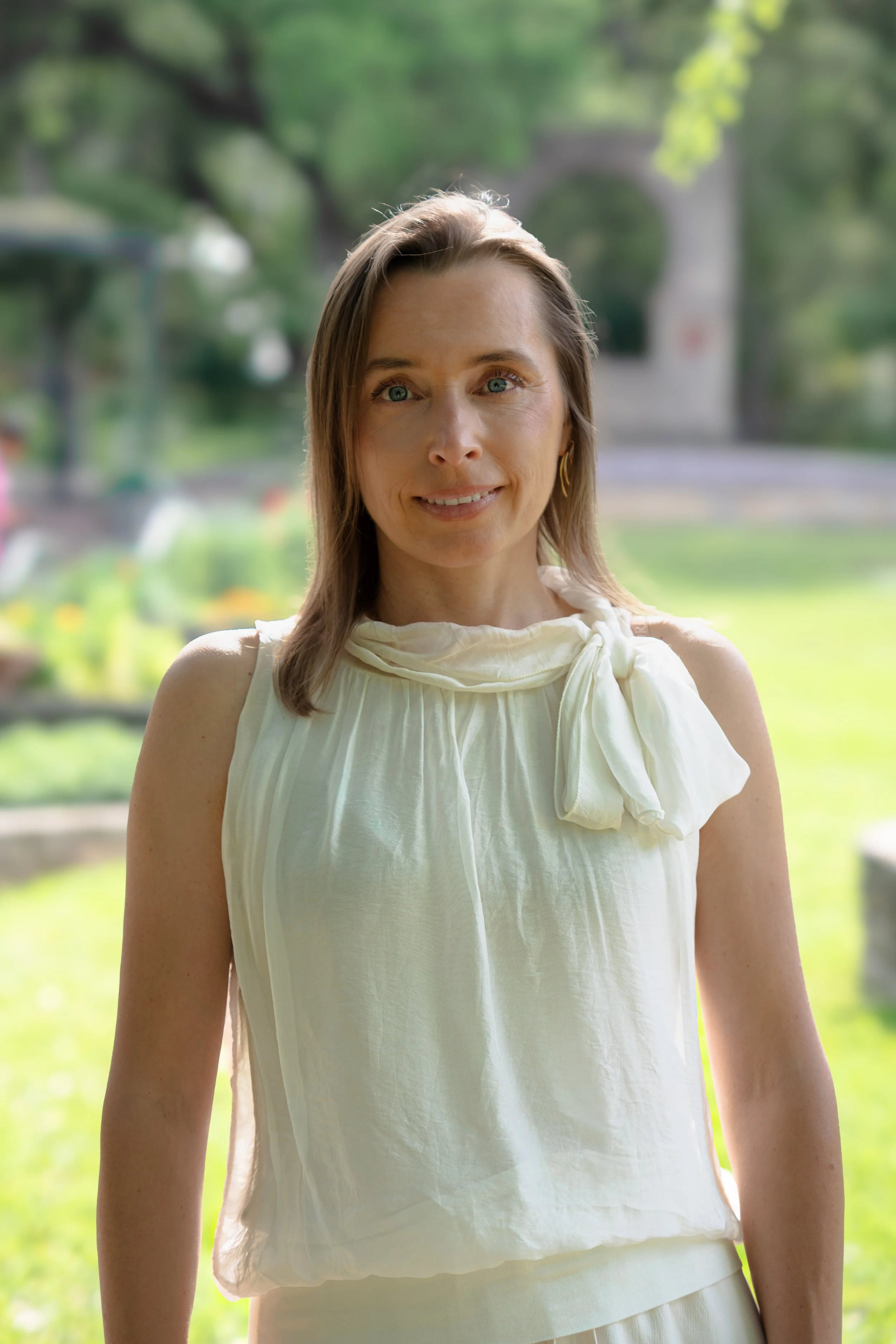 A woman with fair skin and light brown hair standing outdoors in a park or garden with green trees and grass in the background. She is smiling softly and wearing a white sleeveless top with a neckline tied in a bow. The sunlight is softly illuminating her face.