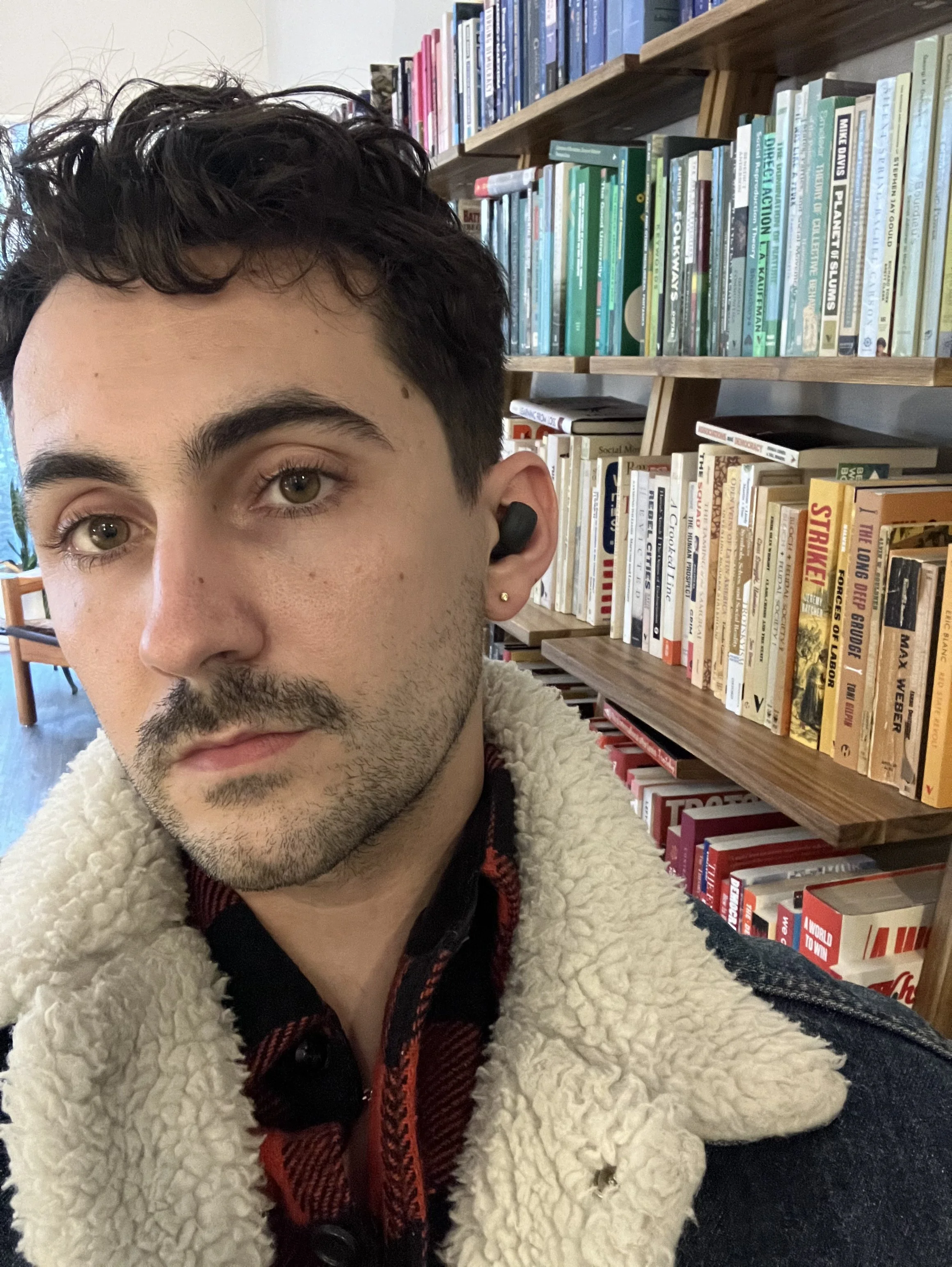 A young man with dark curly hair, brown eyes, and jewelry in his ears, wearing a black and red plaid hoodie under a cream-colored, fluffy fleece jacket, seated in front of bookshelves filled with colorful books in a library or bookstore.