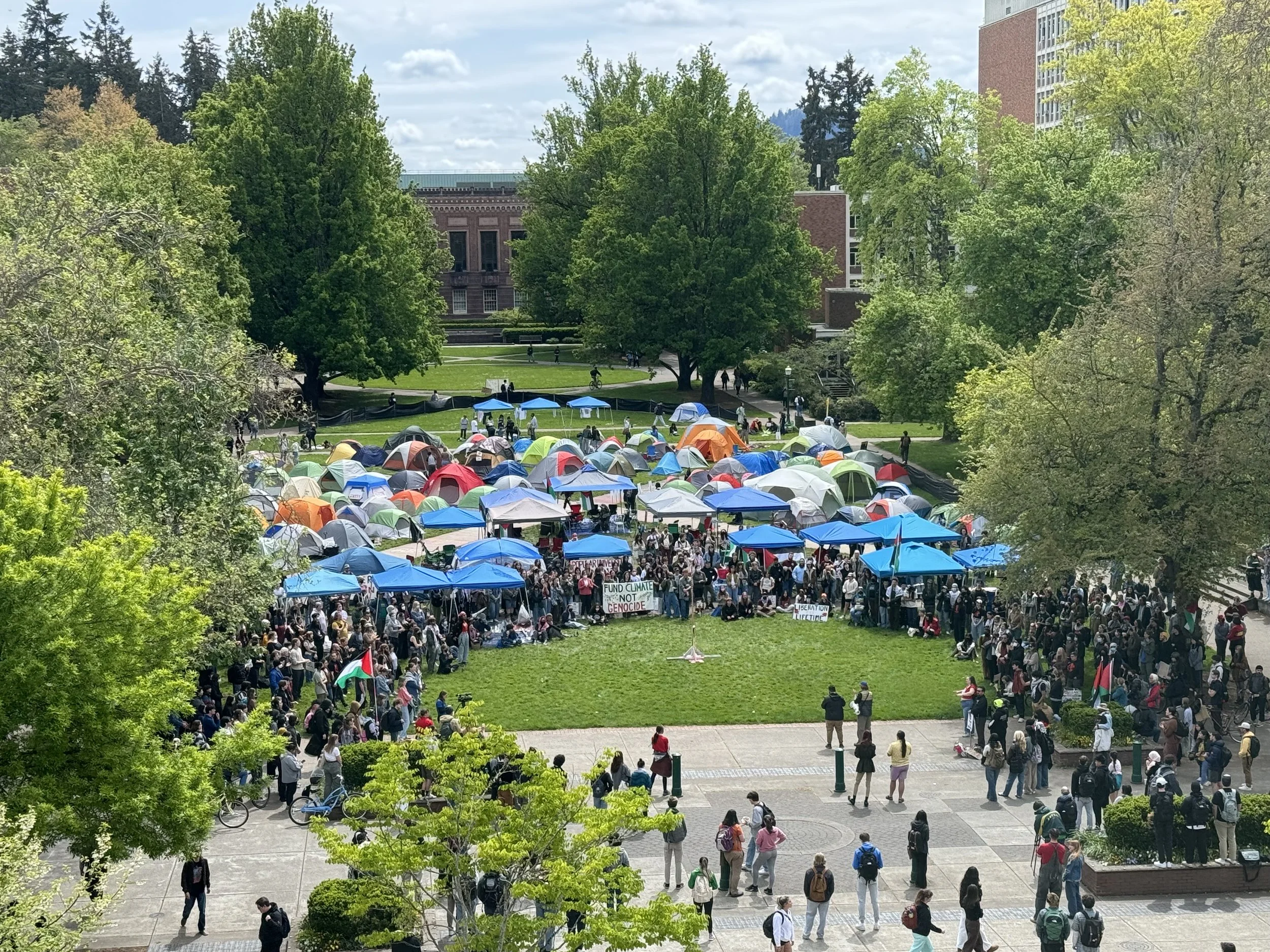 A large crowd of people gathered outdoors in a park with numerous tents, some with signs, during a protest or demonstration. The scene is surrounded by trees and appears to be in an urban area with buildings visible in the background.