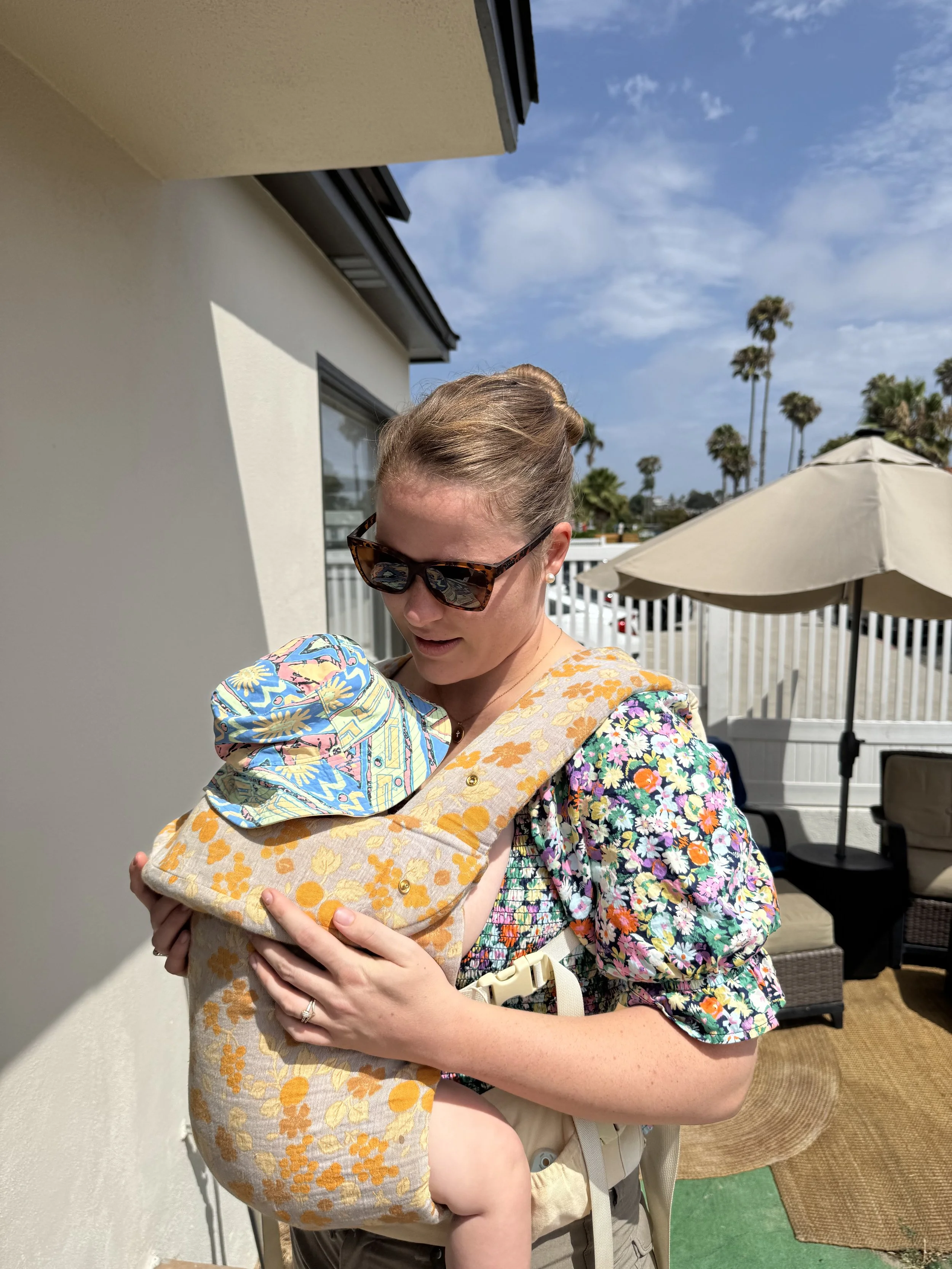 A woman wearing sunglasses and a floral dress is holding a baby. The woman has her hair styled in an updo. The baby is wearing a colorful patterned wrap carrier. They are outdoors under a partly cloudy sky, near patio furniture, including a beige umbrella and chairs, with a background of palm trees.