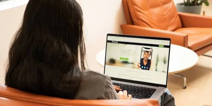 Person with long dark hair using a laptop, sitting on a brown leather chair in a room with a white side table and orange couch.