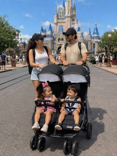 A family with two young children in a double stroller walking towards a castle at a Disney theme park.