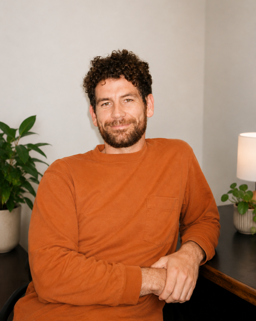 A smiling man with curly hair and beard, wearing an orange long-sleeve shirt, sitting at a desk with a potted plant and a lamp nearby.