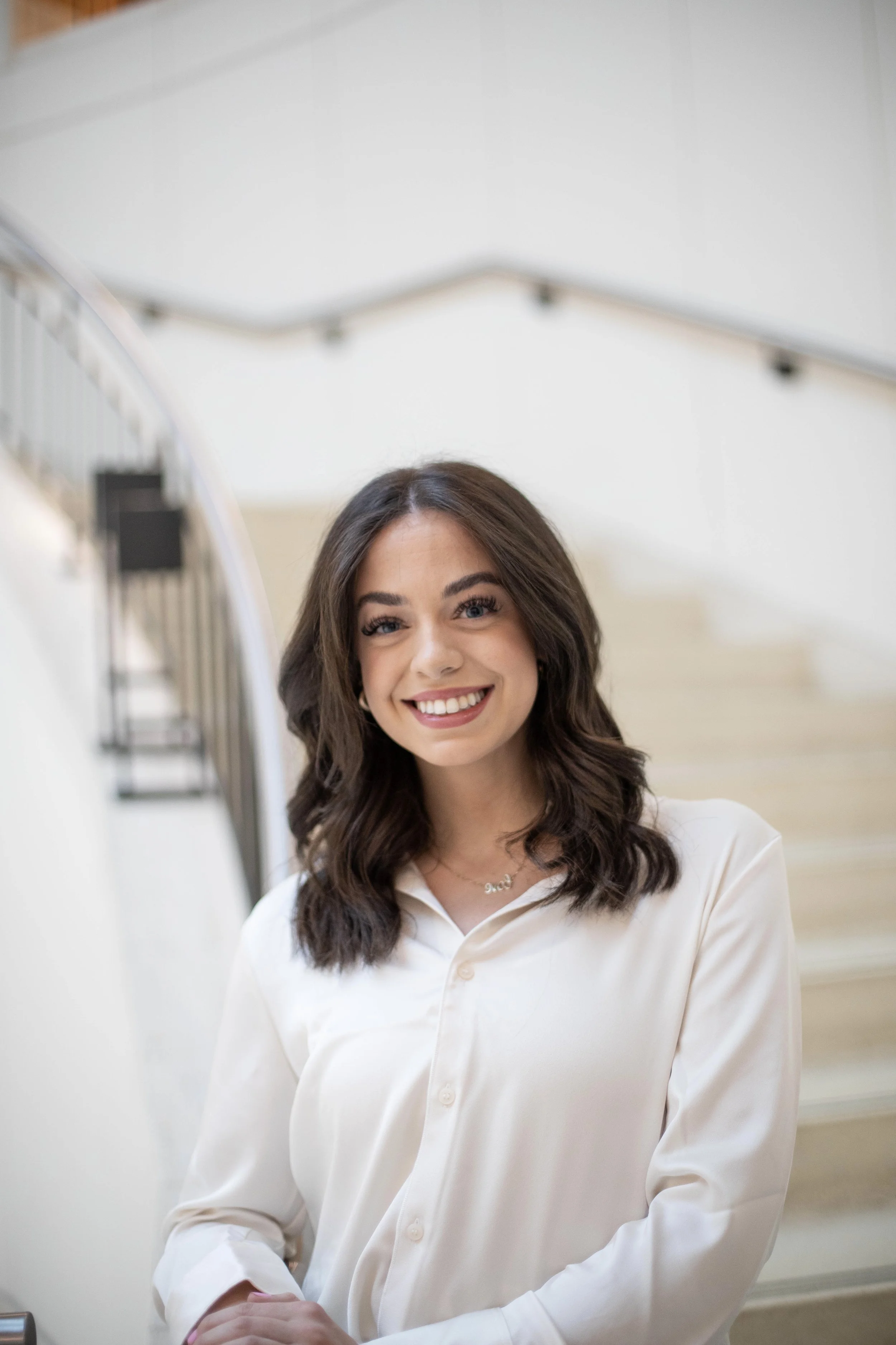 A young woman with brown hair and blue eyes, smiling while standing indoors near a staircase, wearing a white button-down shirt.