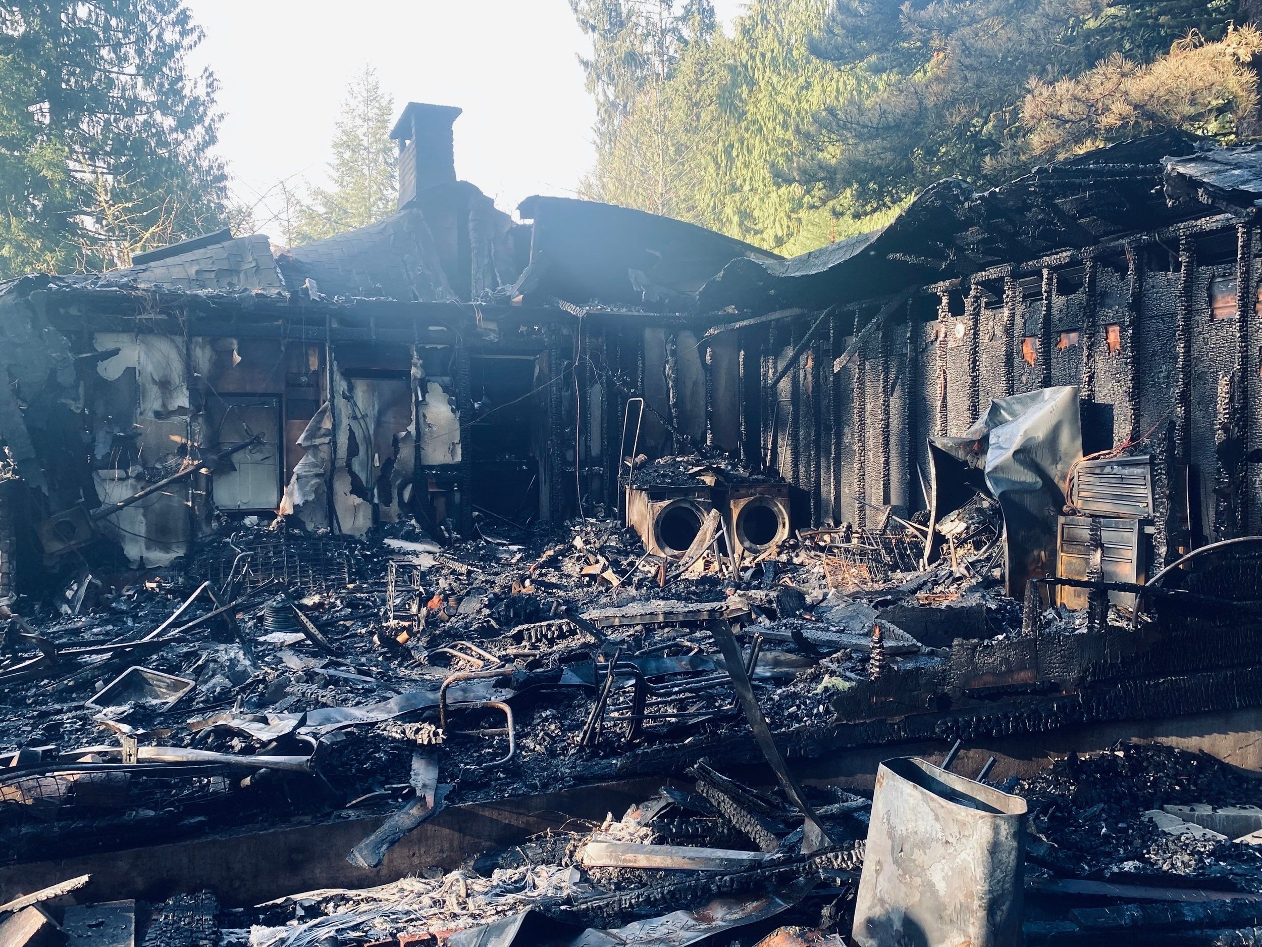 Burned house with charred walls, roof, and scattered debris, surrounded by trees.