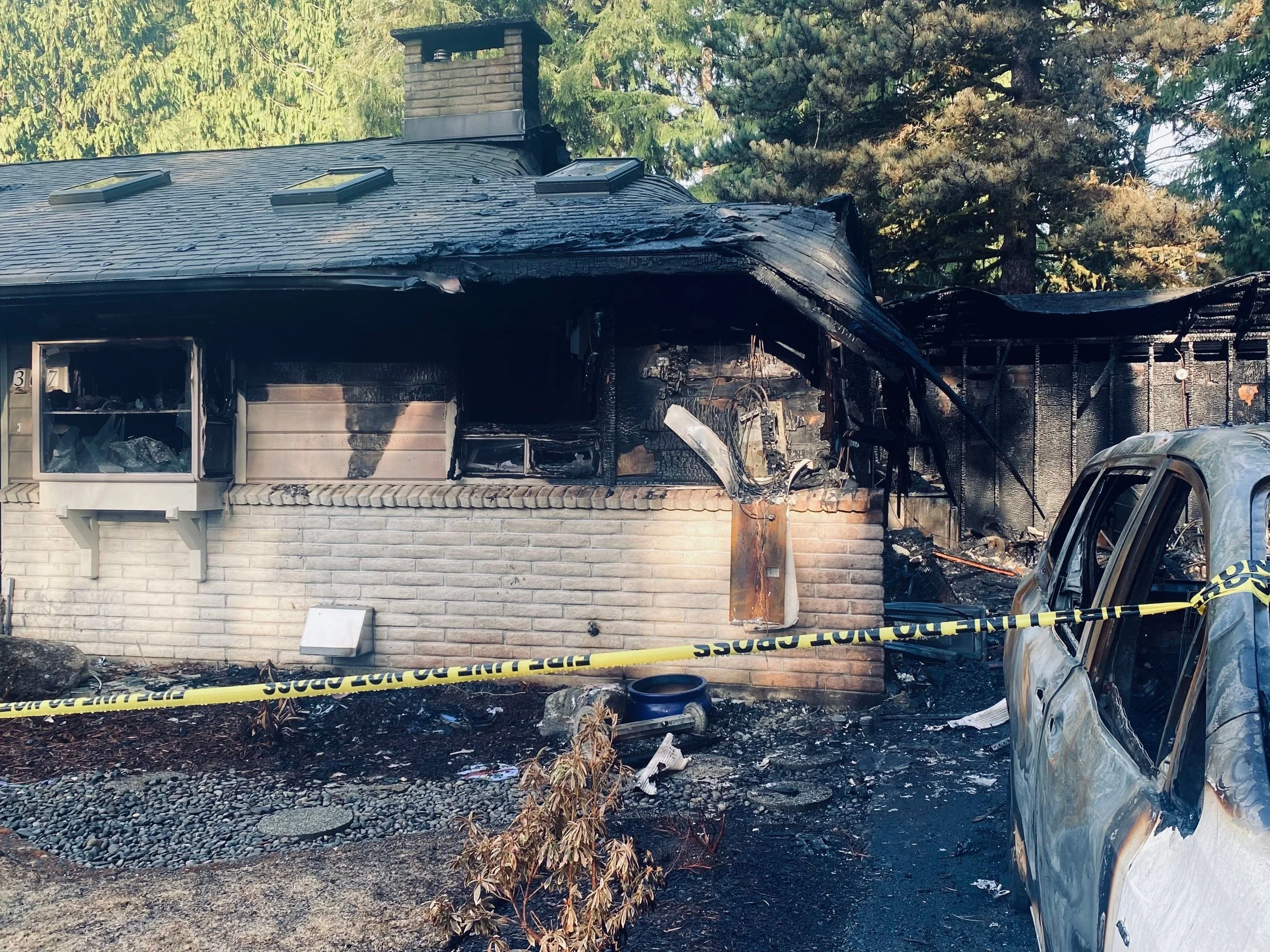 A house with extensive fire damage to the front, showing a burnt and charred roof and walls. A damaged vehicle is parked in front, with caution tape around the scene. Surrounding trees and debris are also visible.