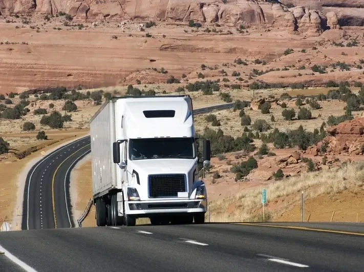 Un camion blanc conduit sur une route sinueuse dans un paysage désertique avec des formations rocheuses rouges.