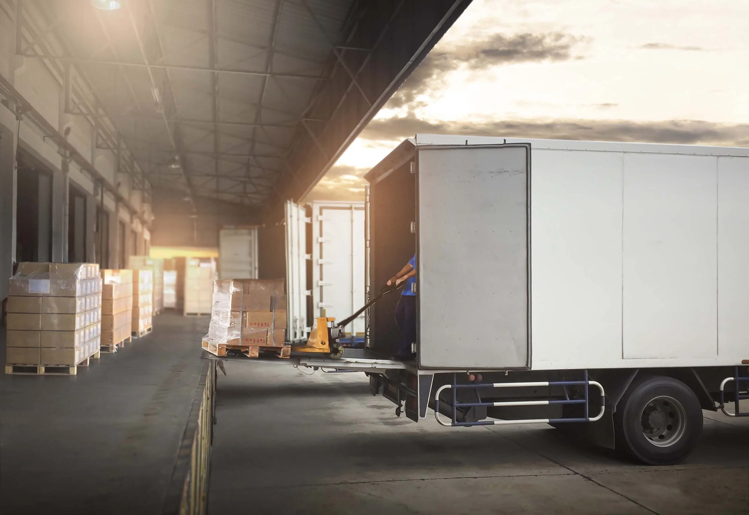 Un camion de livraison stationné dans un entrepôt, avec des caisses en carton sur des palettes à l'arrière, et une personne en train de manipuler le chargement. Le ciel est visible à travers une ouverture dans le bâtiment.