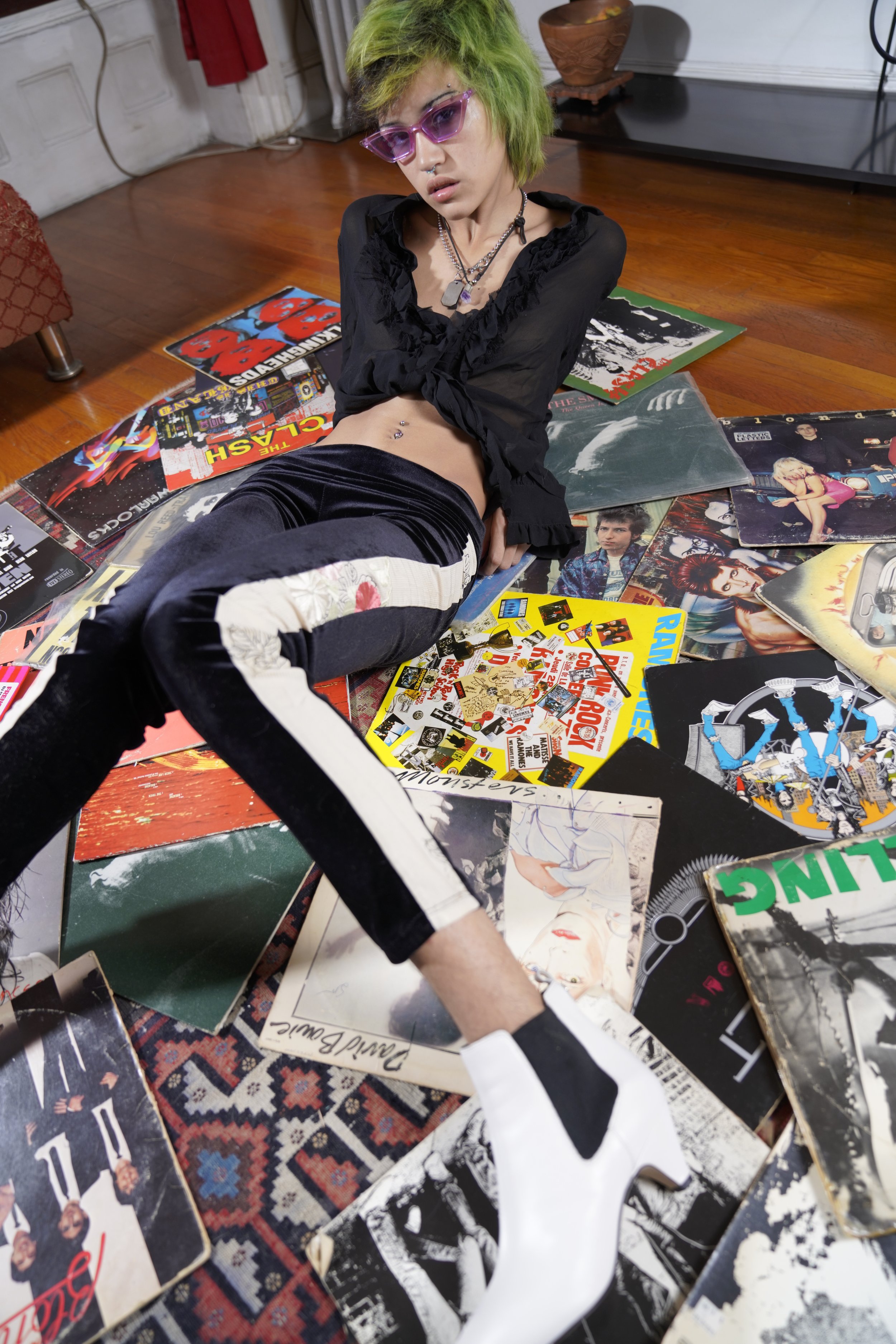 Person with green hair and pink sunglasses sitting on the floor among scattered vinyl record album covers.