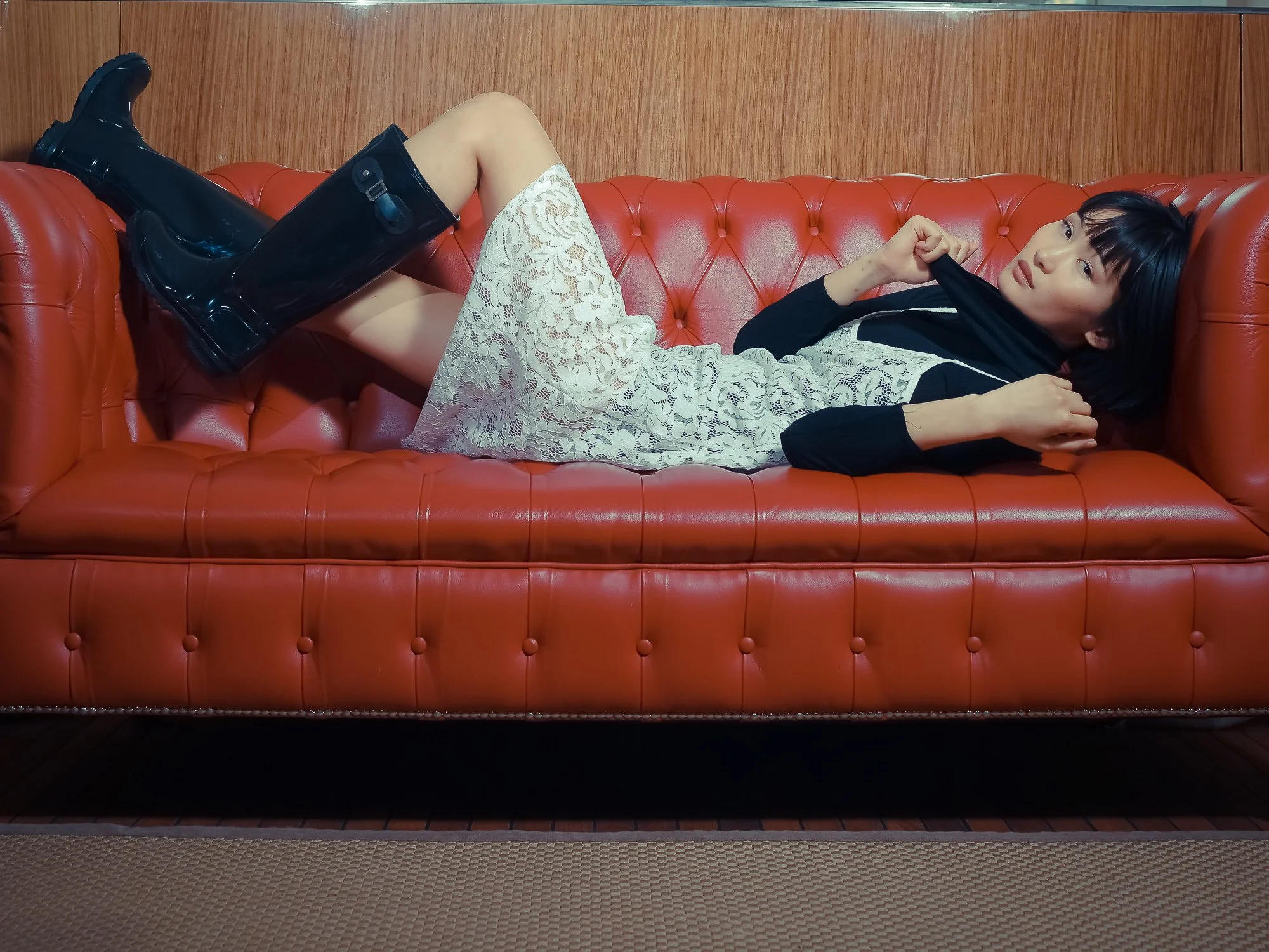 A woman with black hair lying on an orange leather tufted sofa, holding a black piece of clothing near her mouth, wearing a black top, a white lace skirt, and black knee-high rubber boots in a room with wood paneling.