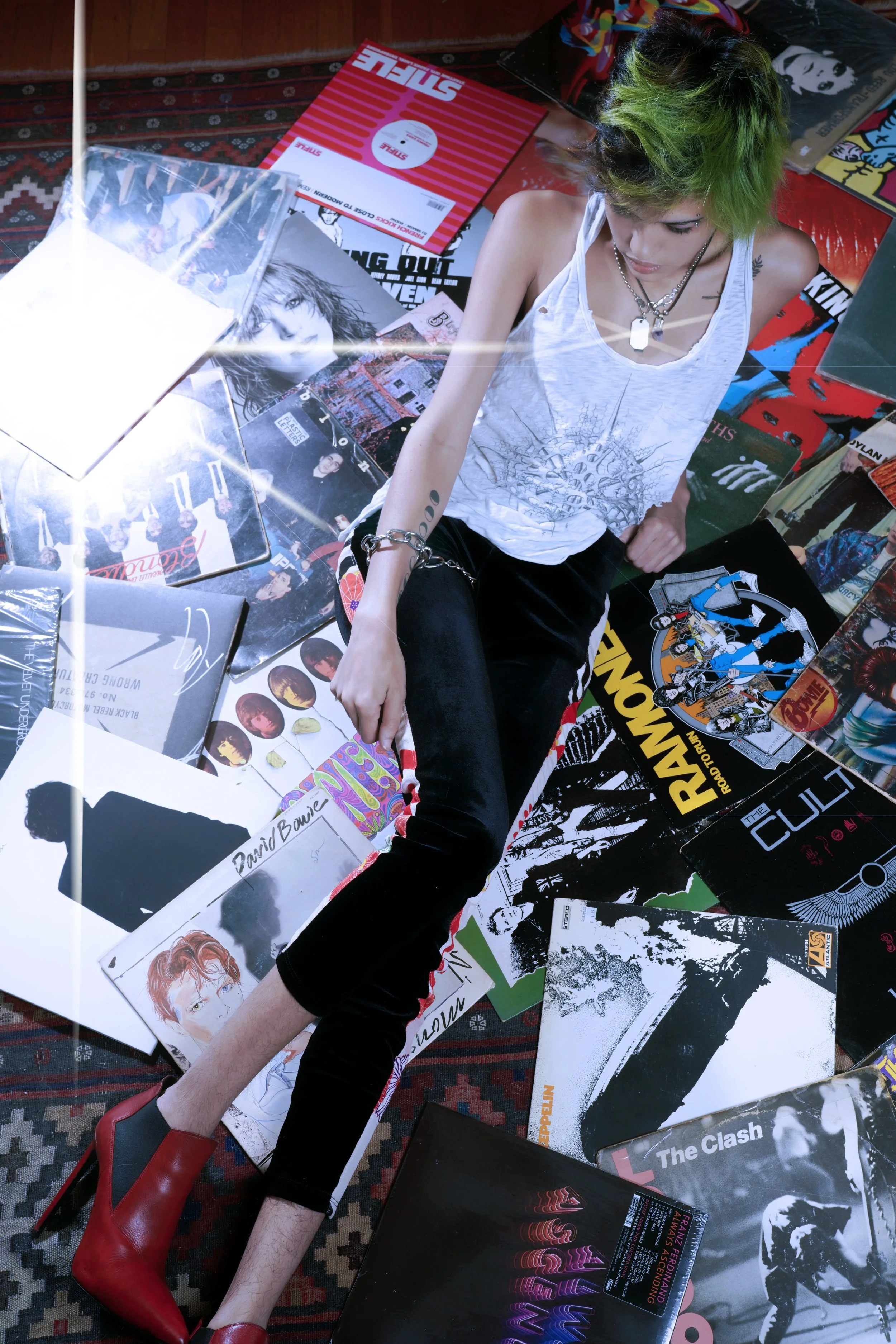 Person with green hair sitting amidst vinyl records and posters on the floor, wearing a white sleeveless shirt, black pants, and red high-heeled boots.