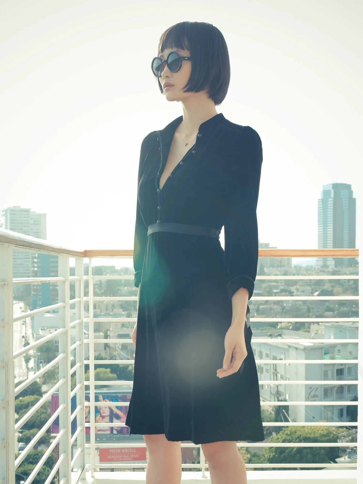 A woman standing on a balcony wearing large black sunglasses and a black dress with buttons down the front, with a cityscape and tall buildings in the background during daytime.