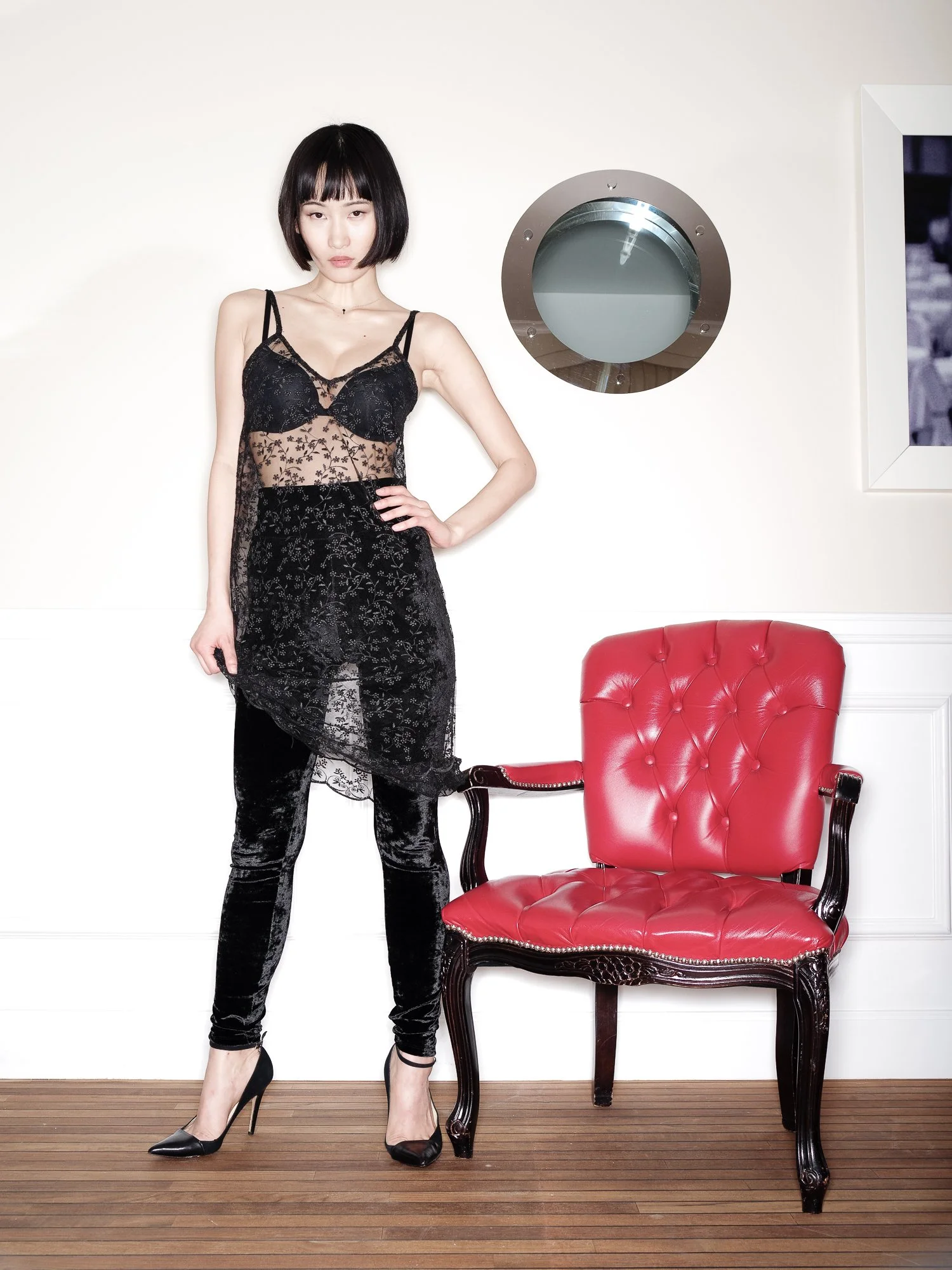 A woman with short black hair and bangs, wearing a black lace dress and high heels, standing with one hand on her hip, next to a red upholstered chair, in a room with white walls and wooden flooring.