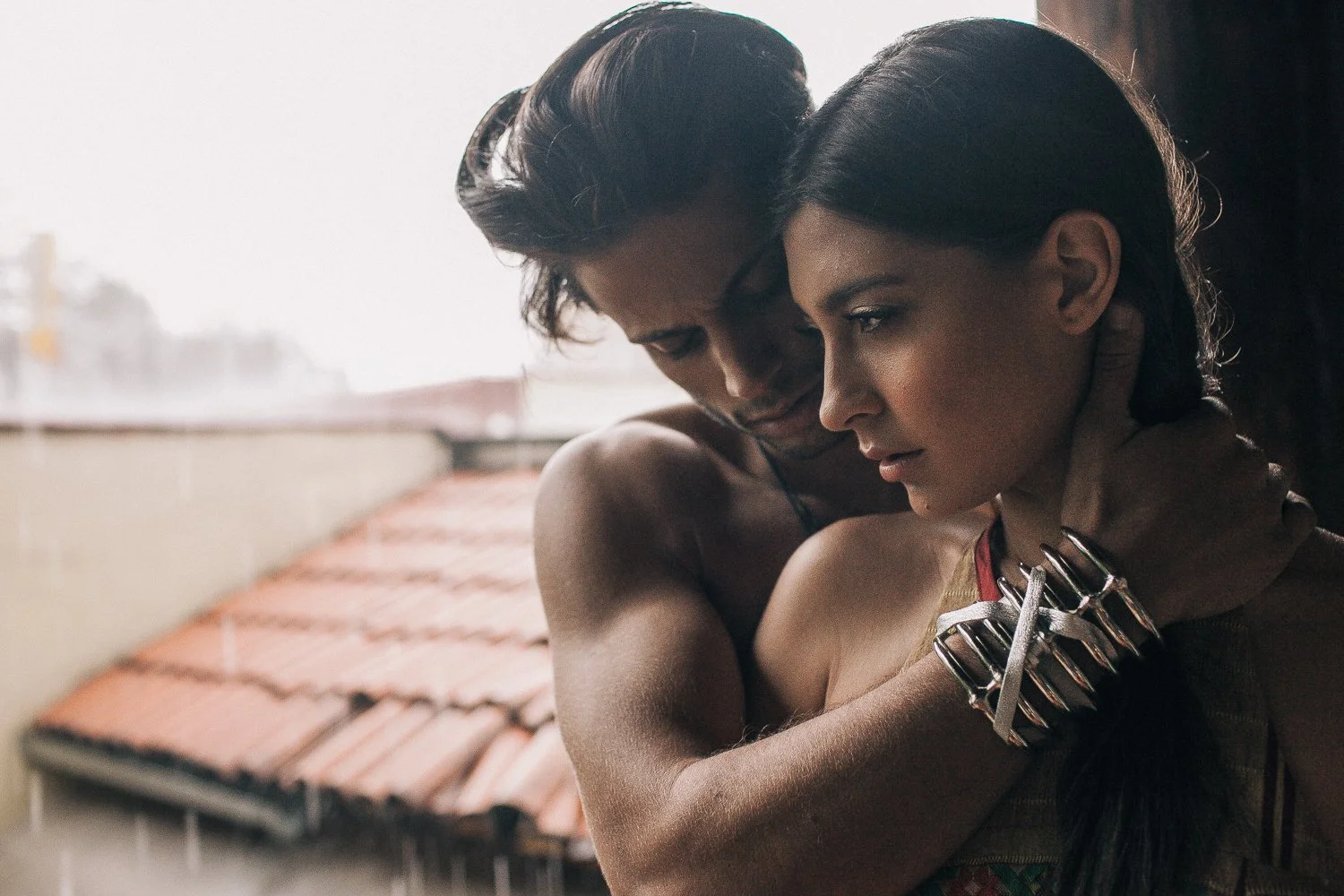 A man and woman embrace closely, with their foreheads touching and eyes closed, outdoors near a building with a tiled roof.