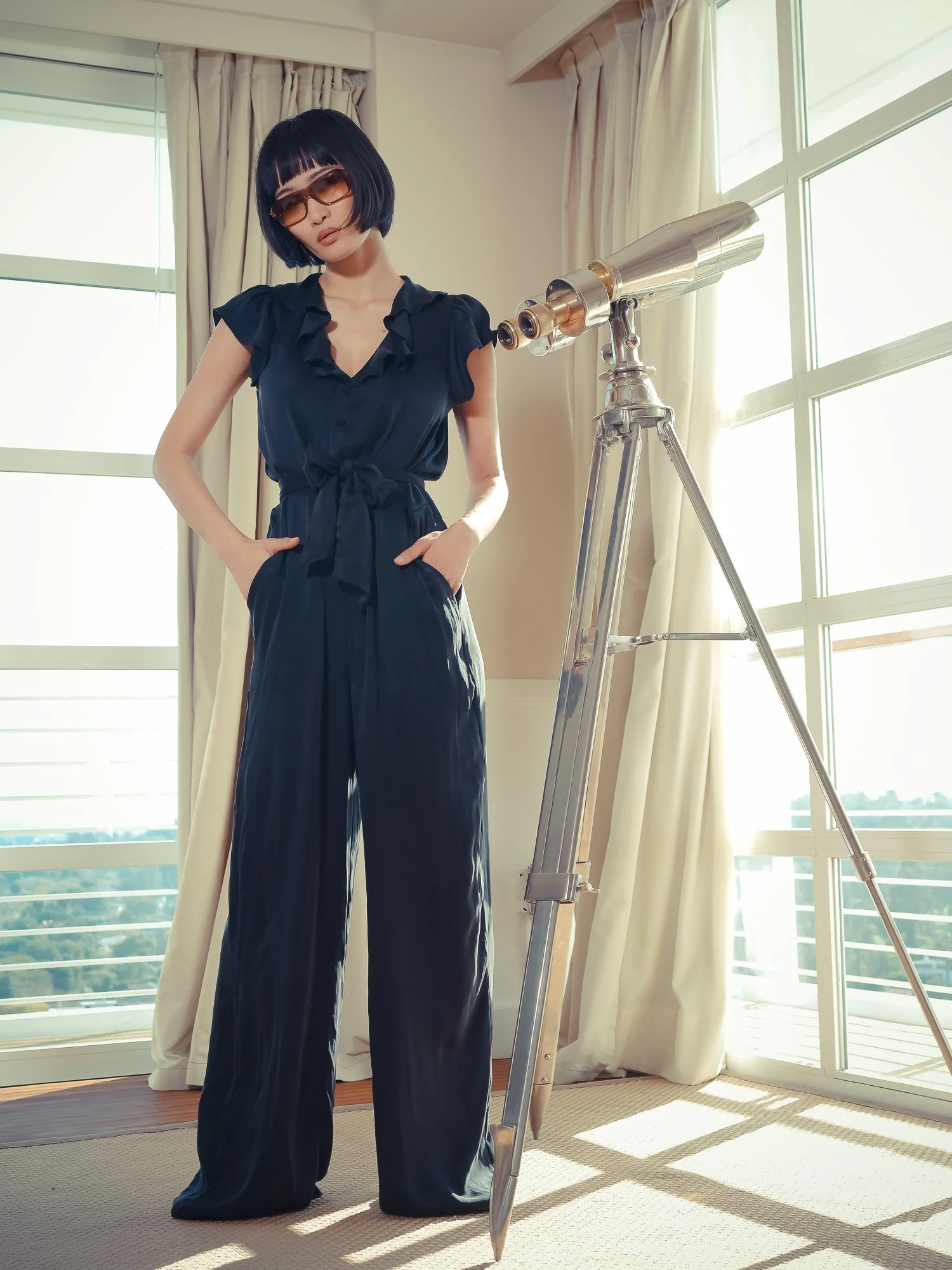 A woman with short black hair, wearing sunglasses and a dark blue dress, stands indoors near a telescope by large windows with sunlight streaming in.