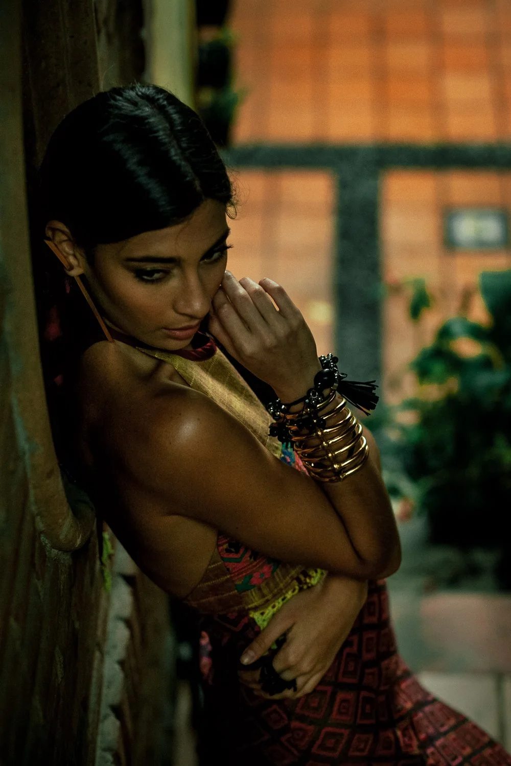 A woman with dark hair and makeup, wearing colorful patterned clothing and multiple gold and black bracelets, leaning against a wood wall with a blurred background of an indoor setting with plants.