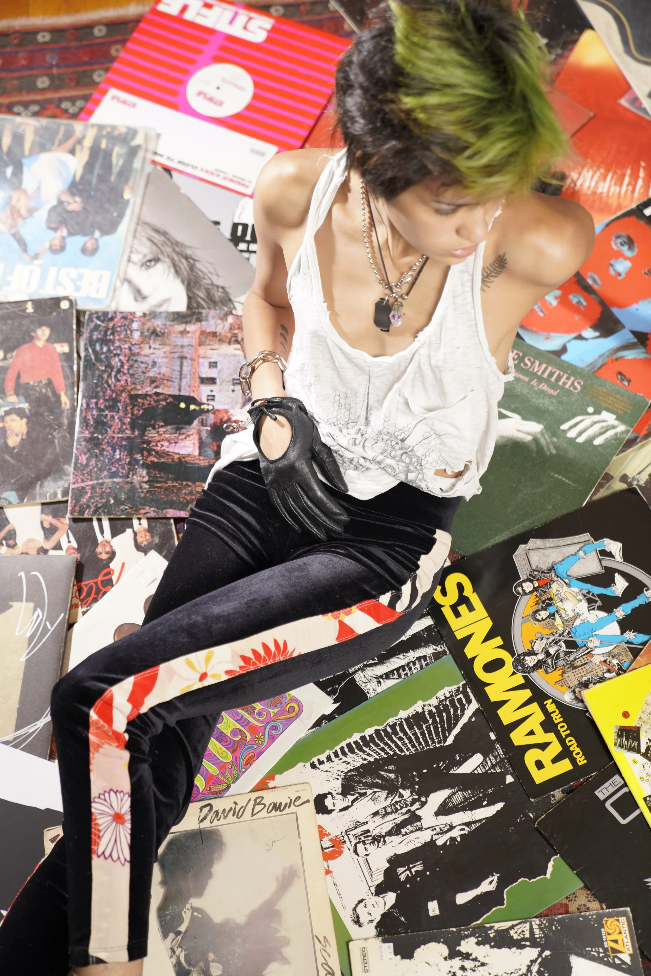 A person with green and black hair lying on the floor surrounded by vinyl records and album covers, wearing a ripped white tank top, black pants with a floral strip on the side, earrings, necklaces, and a black glove on one hand.