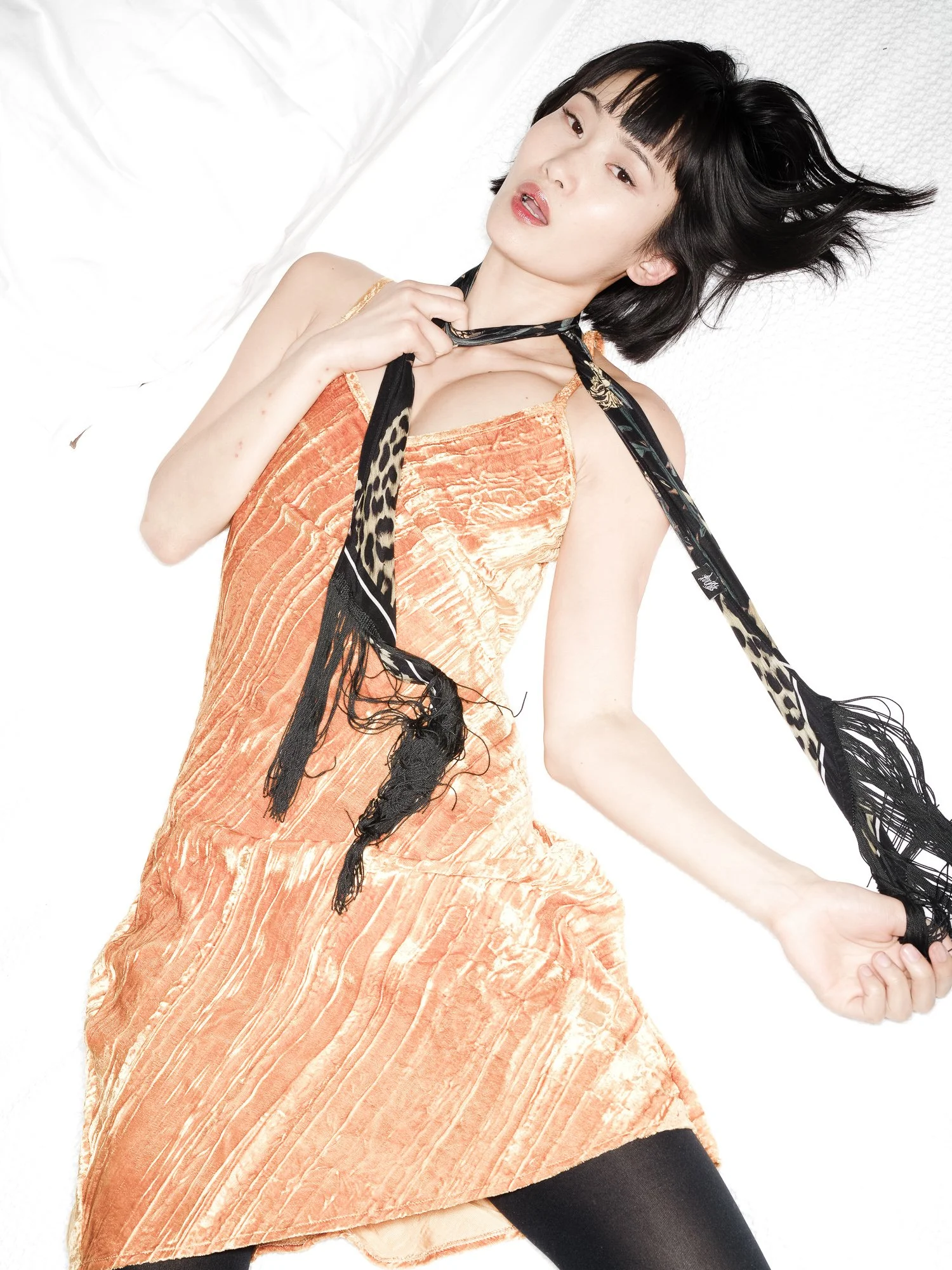 A woman lying on a white surface, wearing an orange dress and black tights, holding a black and animal print scarf around her neck, with her hair styled in a short, black bob