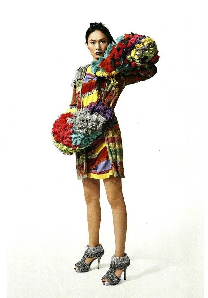 Woman wearing a colorful, patterned dress and high-heeled shoes, holding large, multicolored fluffy objects.