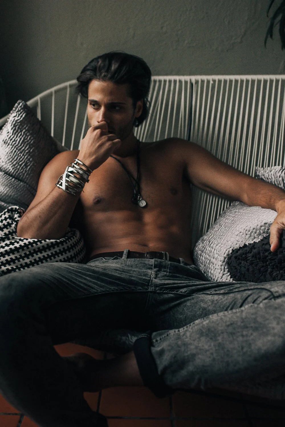 A shirtless young man with dark hair, wearing multiple bracelets and a necklace, lies on a striped sofa with patterned pillows, in a dimly lit room, resting his head on one hand and looking thoughtfully to the side.