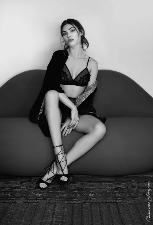 A woman sitting on a sofa in lingerie, with a black jacket draped over her shoulders, in black and white photo.