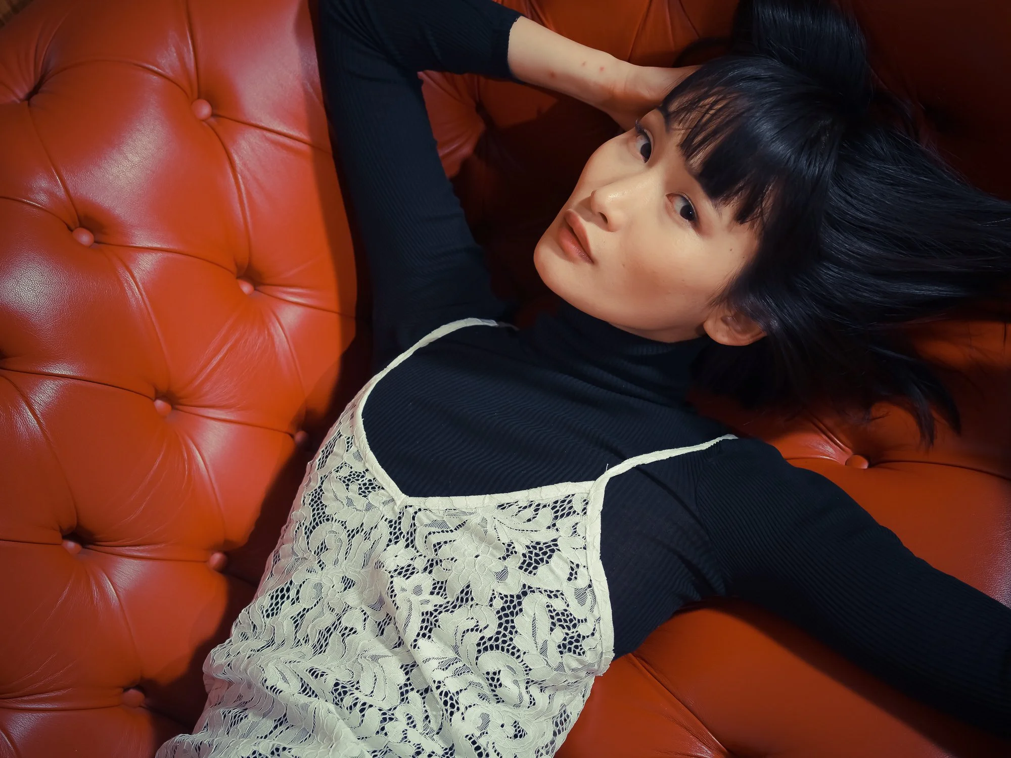 Young woman with dark hair and bangs lying on a caramel-colored leather couch, wearing a black top and a beige lace apron.
