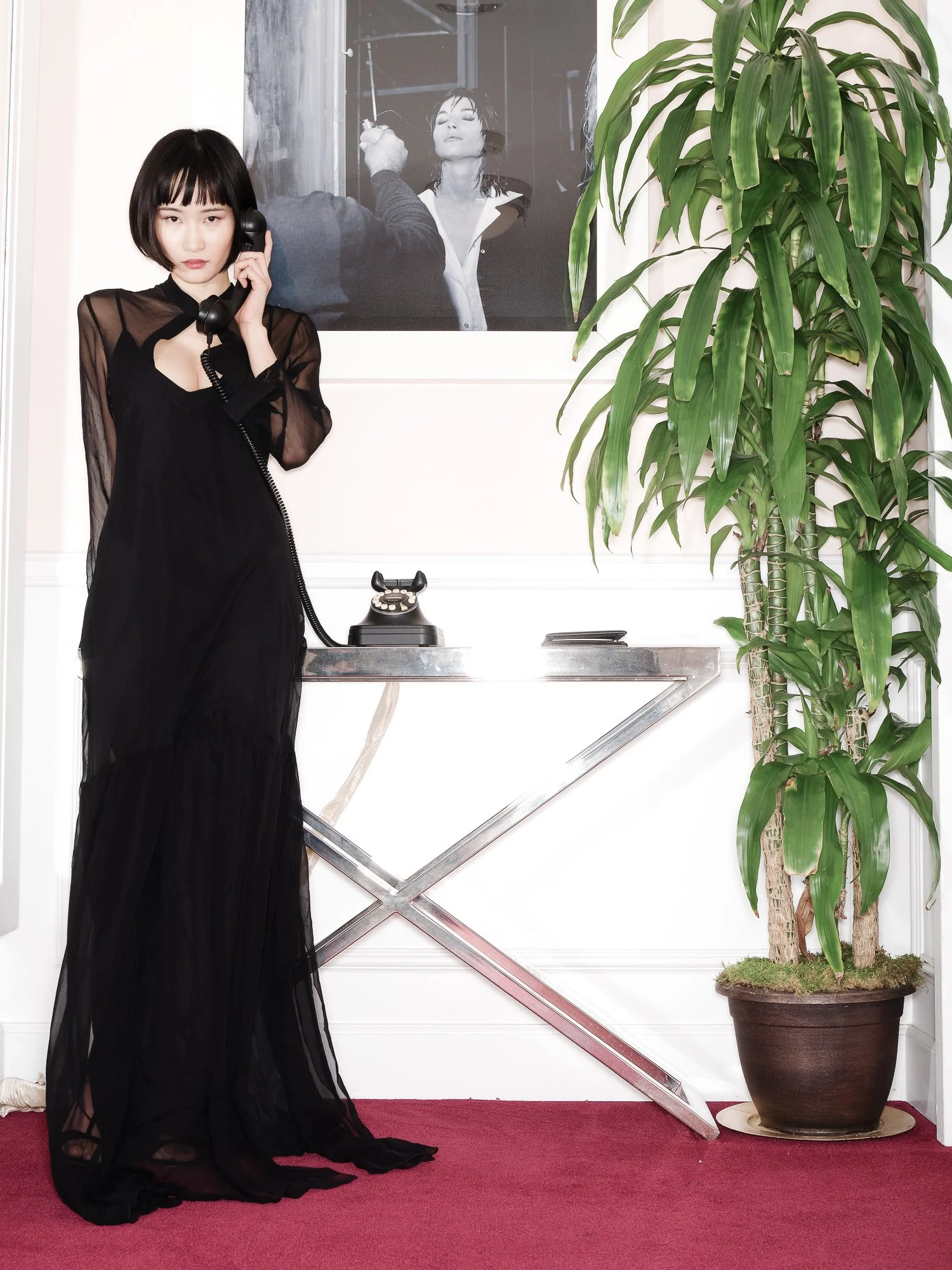 A woman with short black hair and a black dress with a sheer top is holding a vintage black rotary telephone to her ear while standing on a red carpet in front of a white wall with a large potted plant on the right and a framed black and white photo 