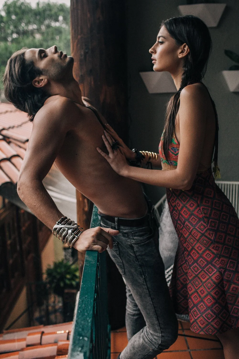 A shirtless man with long dark hair leaning back on a balcony railing, and a woman with long dark hair in a braid wearing a colorful dress, standing close to him with her hand on his chest, facing each other indoors during daytime.