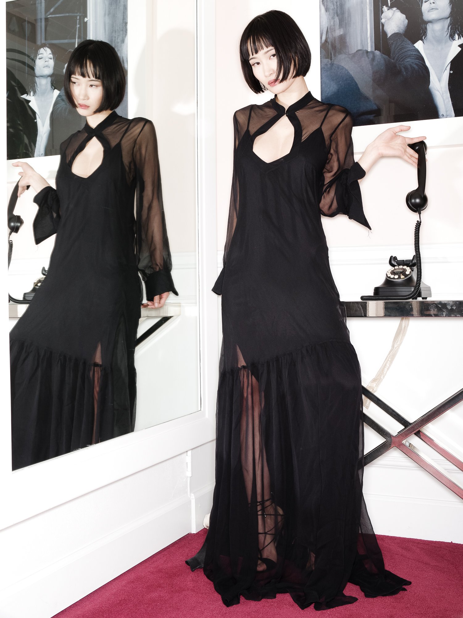 A woman in a sheer black dress with ruffled hem, standing on a red carpet next to a mirror with black and white photographs in the background. She holds a telephone receiver in one hand and has a bob haircut with bangs.