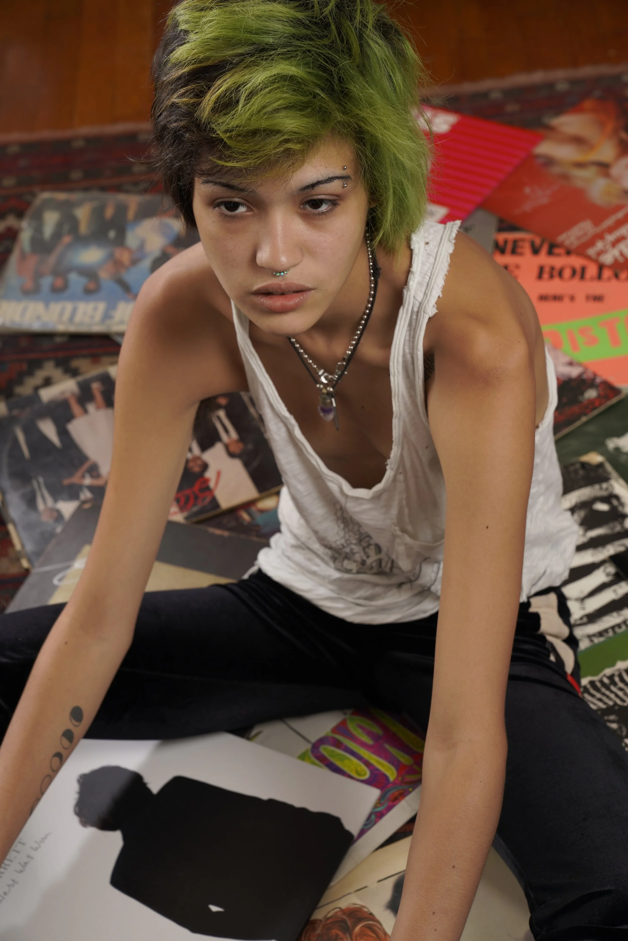 Young person with green and black hair, pierced nose and eyebrow, wearing white ripped tank top and layered necklaces, sitting on the floor with posters and artwork around.