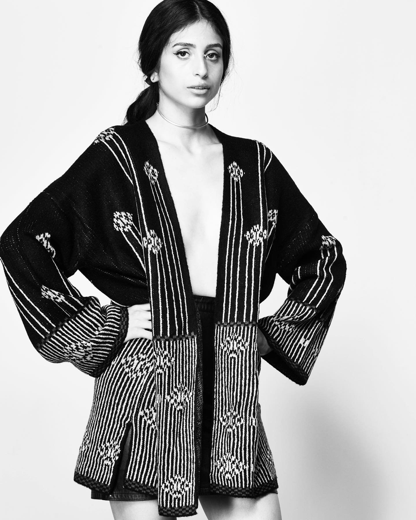 Black and white photo of a woman with dark hair in a loose ponytail, wearing a patterned, embroidered cardigan and black shorts, standing against a plain light background.