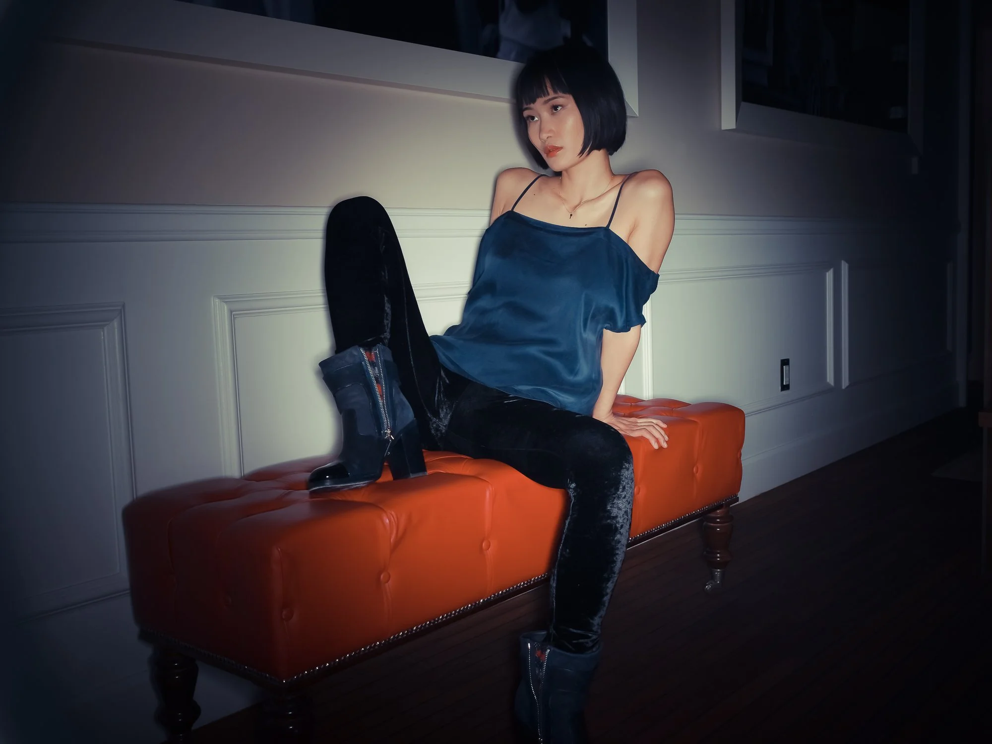 A woman with short black hair and bangs is sitting on an orange tufted bench against a white paneled wall. She is wearing a blue satin camisole, black velvet pants, and black platform boots. She is posing with one leg up on the bench, looking to her 