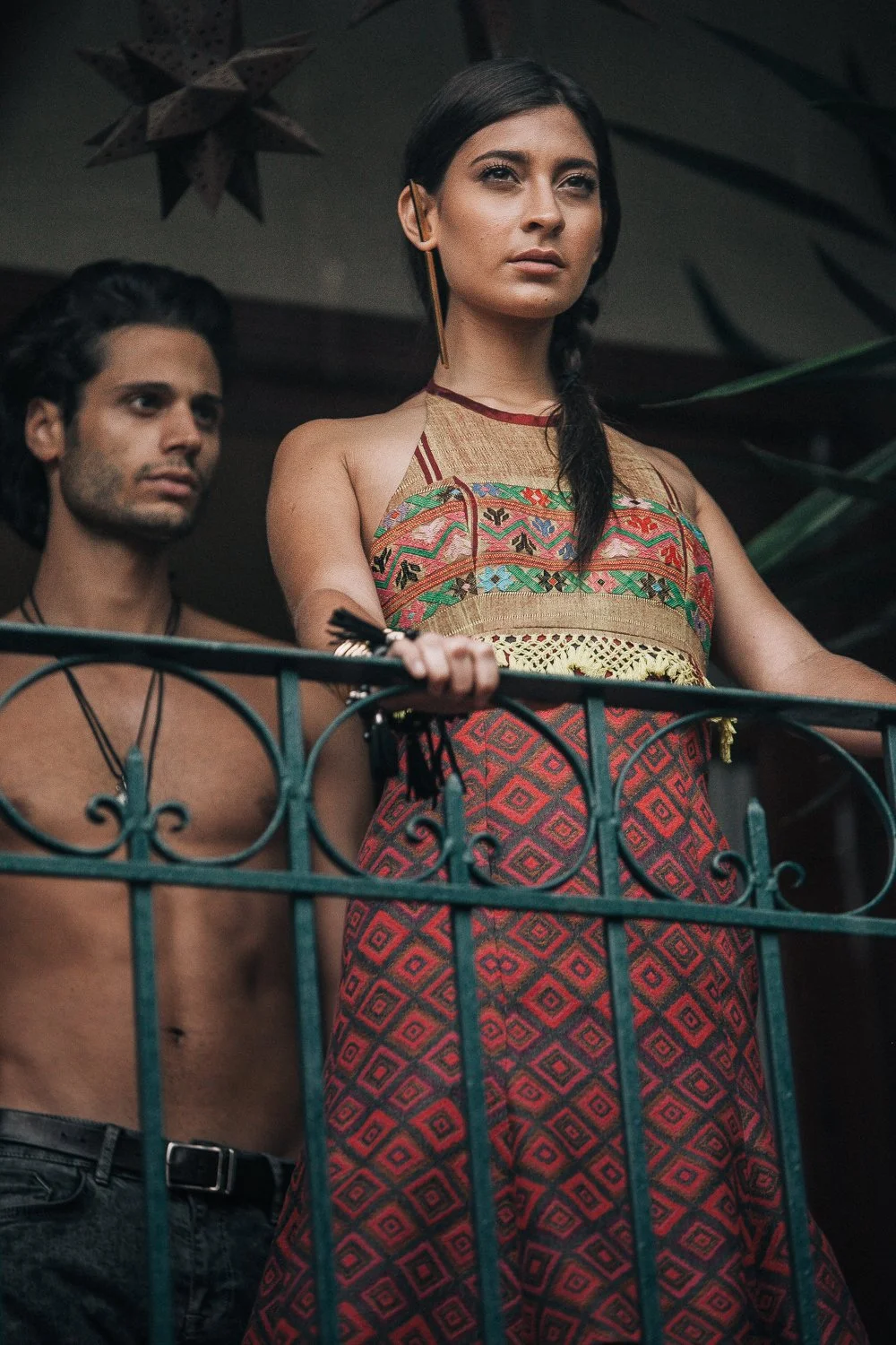 A woman with long dark hair and tan skin, wearing a colorful embroidered sleeveless top and a red patterned skirt, stands behind a teal metal railing, with a shirtless man with medium skin tone and dark hair standing behind her.