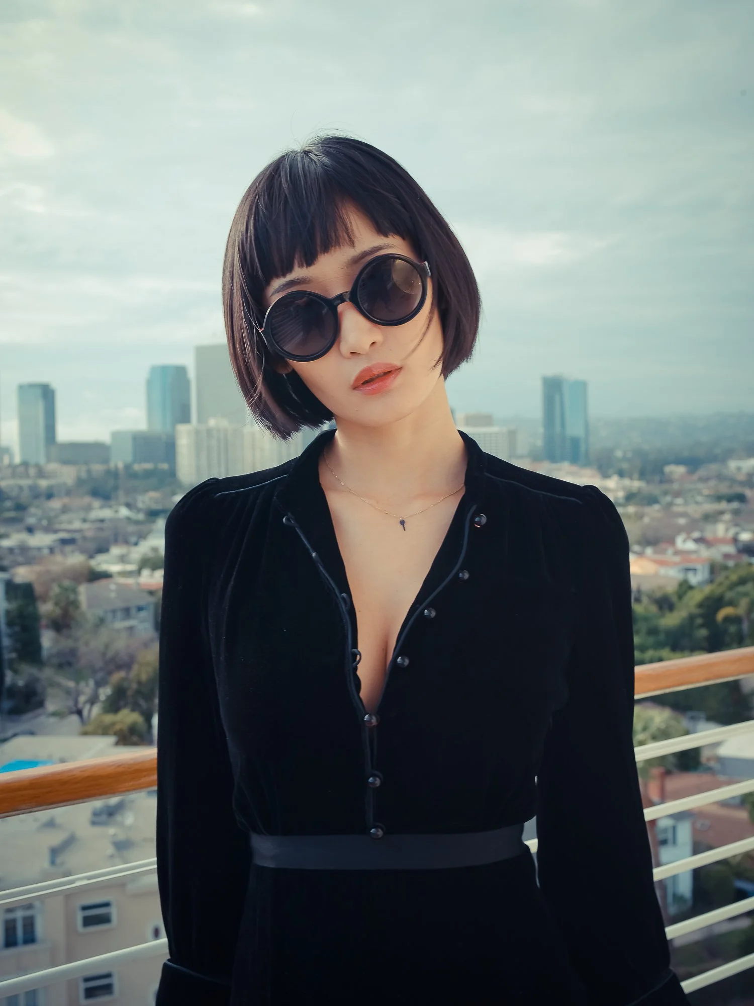 A woman with short black hair wearing large round sunglasses and a black top standing outdoors with a cityscape in the background.