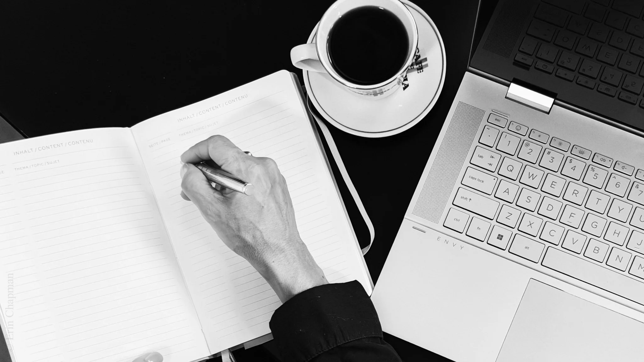 Top view of a person writing in an open notebook beside a laptop and coffee cup