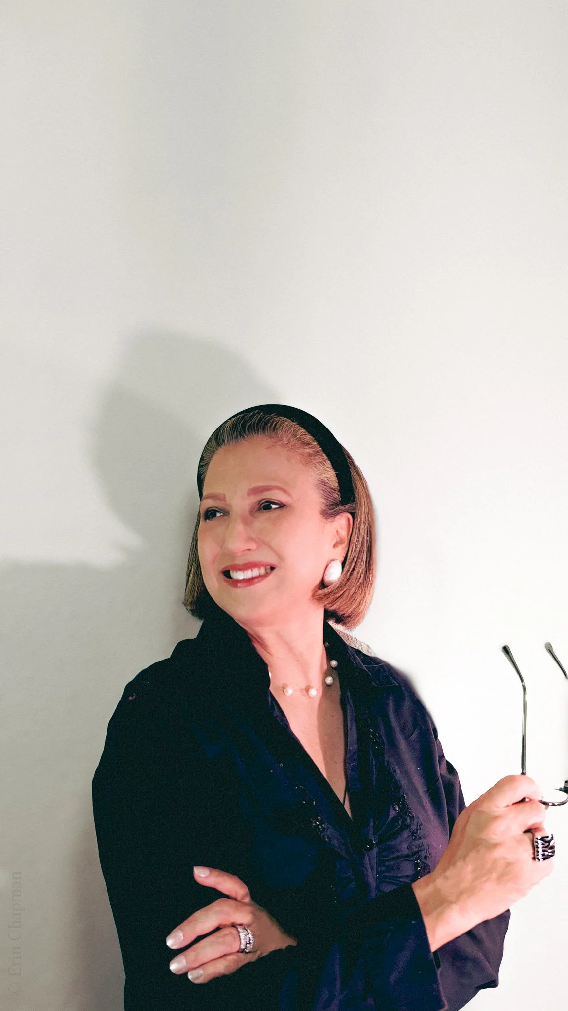 Smiling woman in a black blouse holding eyeglasses against a white wall