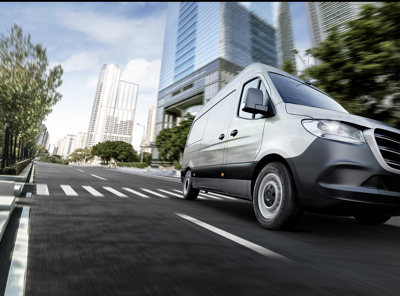 A white delivery van driving through a city street with tall modern buildings and green trees on a sunny day.