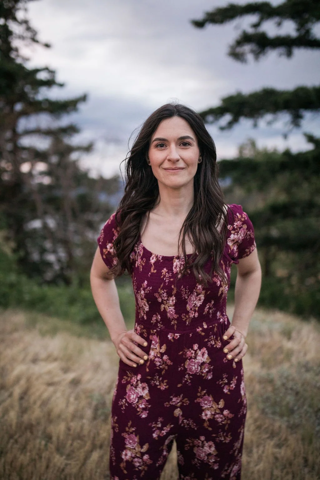 A young woman with long dark hair standing outdoors in a grassy area with trees and a cloudy sky in the background. She is wearing a maroon floral jumpsuit and has her hands on her hips, smiling softly.
