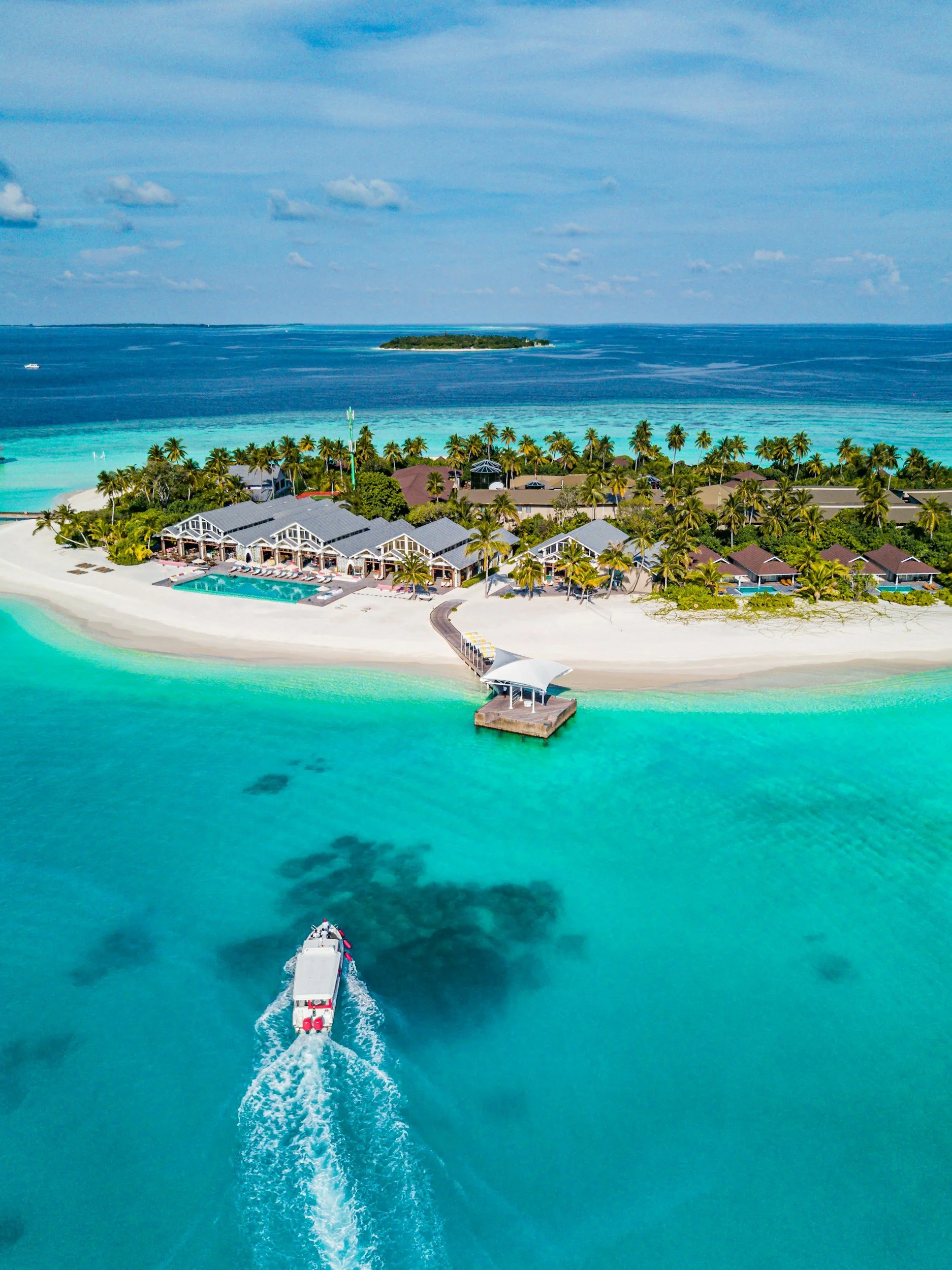 Aerial view of a tropical island resort with buildings, palm trees, a boat approaching a dock, turquoise water, and an ocean in the background.