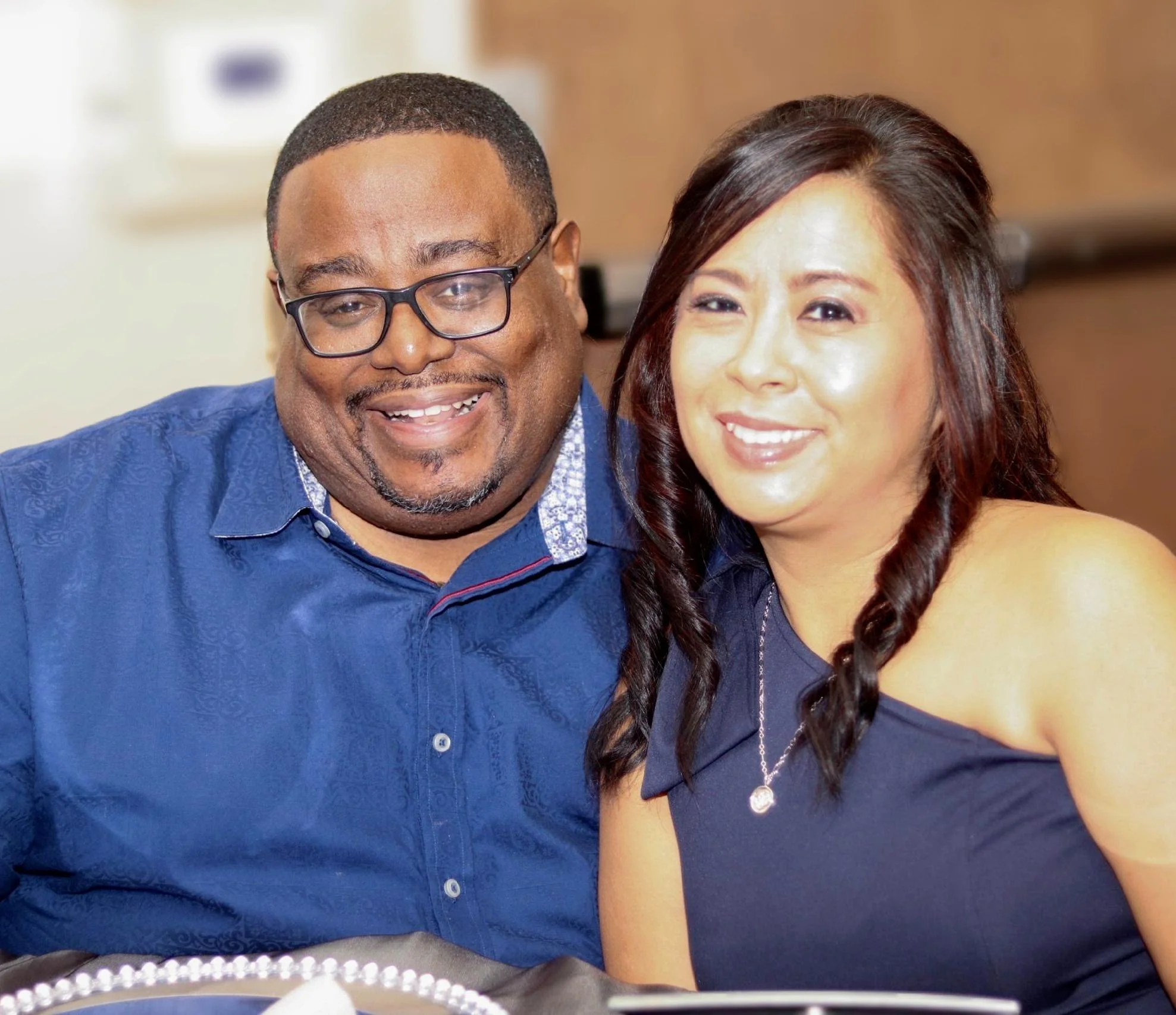 Close-up of a smiling couple, a man with glasses and a woman with dark hair, indoors with a blurred background.