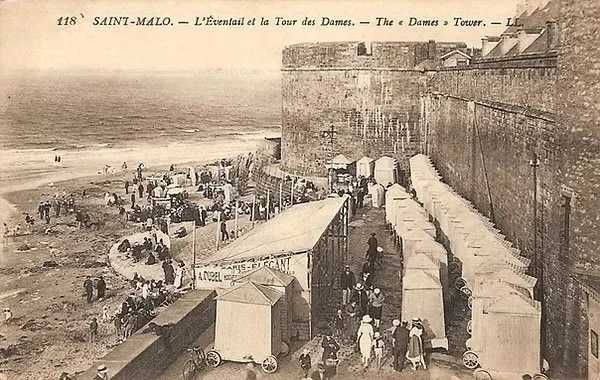 A historical black-and-white photo of the Eiffel Tower and the Seine River at Saint-Malo, showing a busy scene with tents, market stalls, and people walking along a promenade.