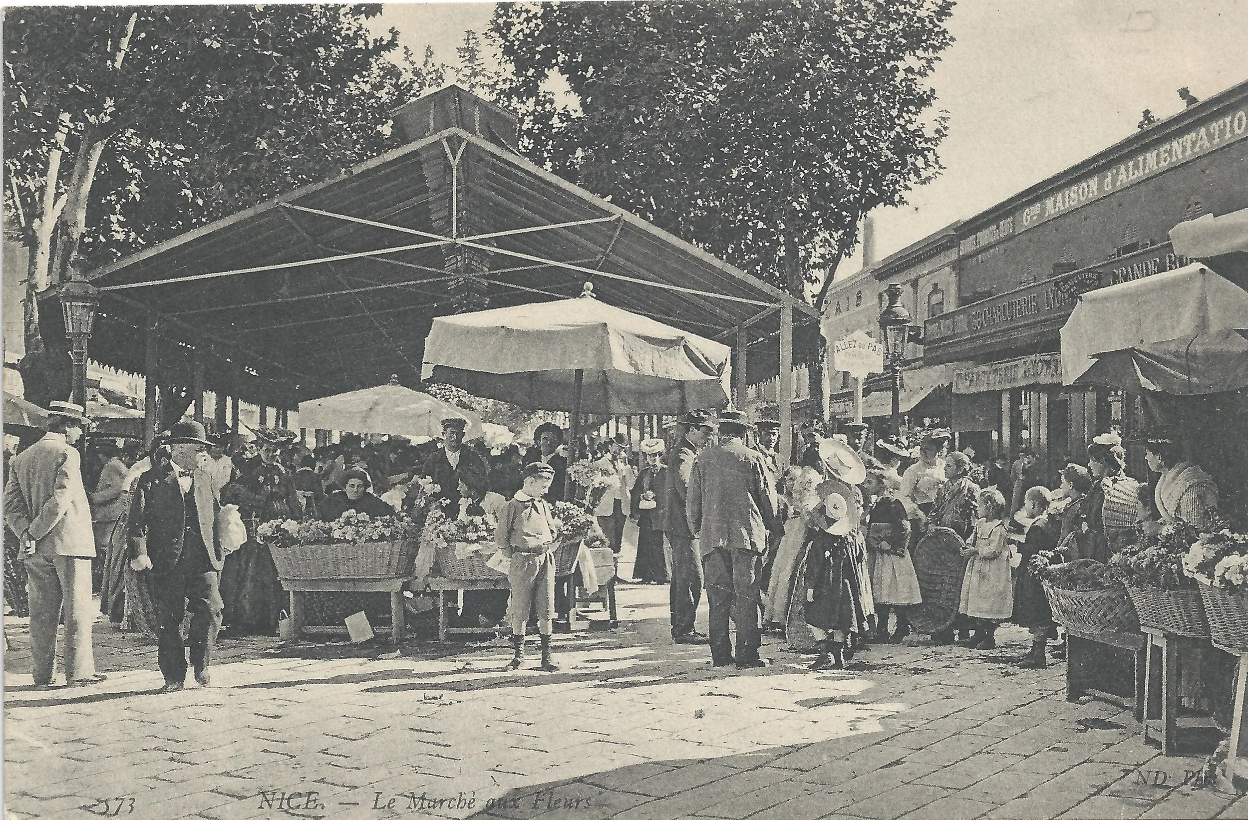 A black and white vintage photograph of a busy outdoor marketplace with numerous people shopping for flowers and goods. There are market stalls with umbrellas, large trees providing shade, and some buildings in the background with signs in French.