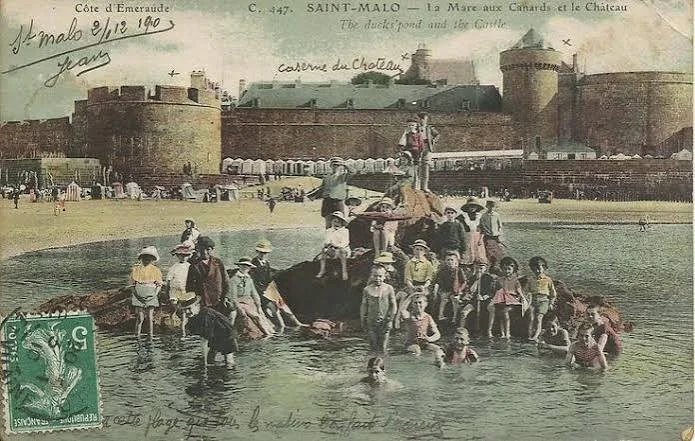 Historical colorized photo of a large group of people, including children, bathing and swimming in a river near a fortress called Château in Saint-Malo, France. The fortress wall and towers are visible in the background.
