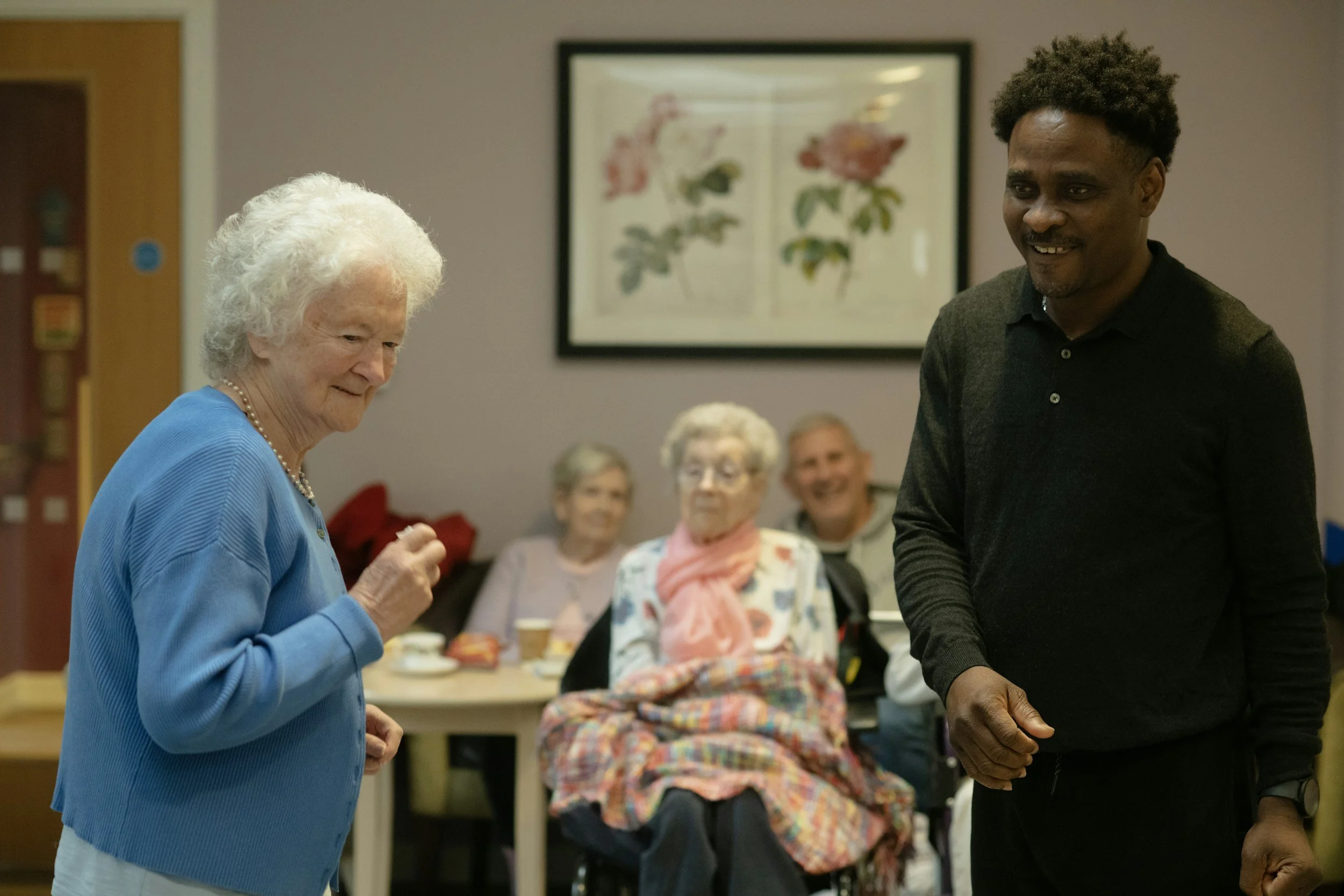 An elderly woman and a young man smiling at each other in a social gathering room, with three other elderly women seated behind them.