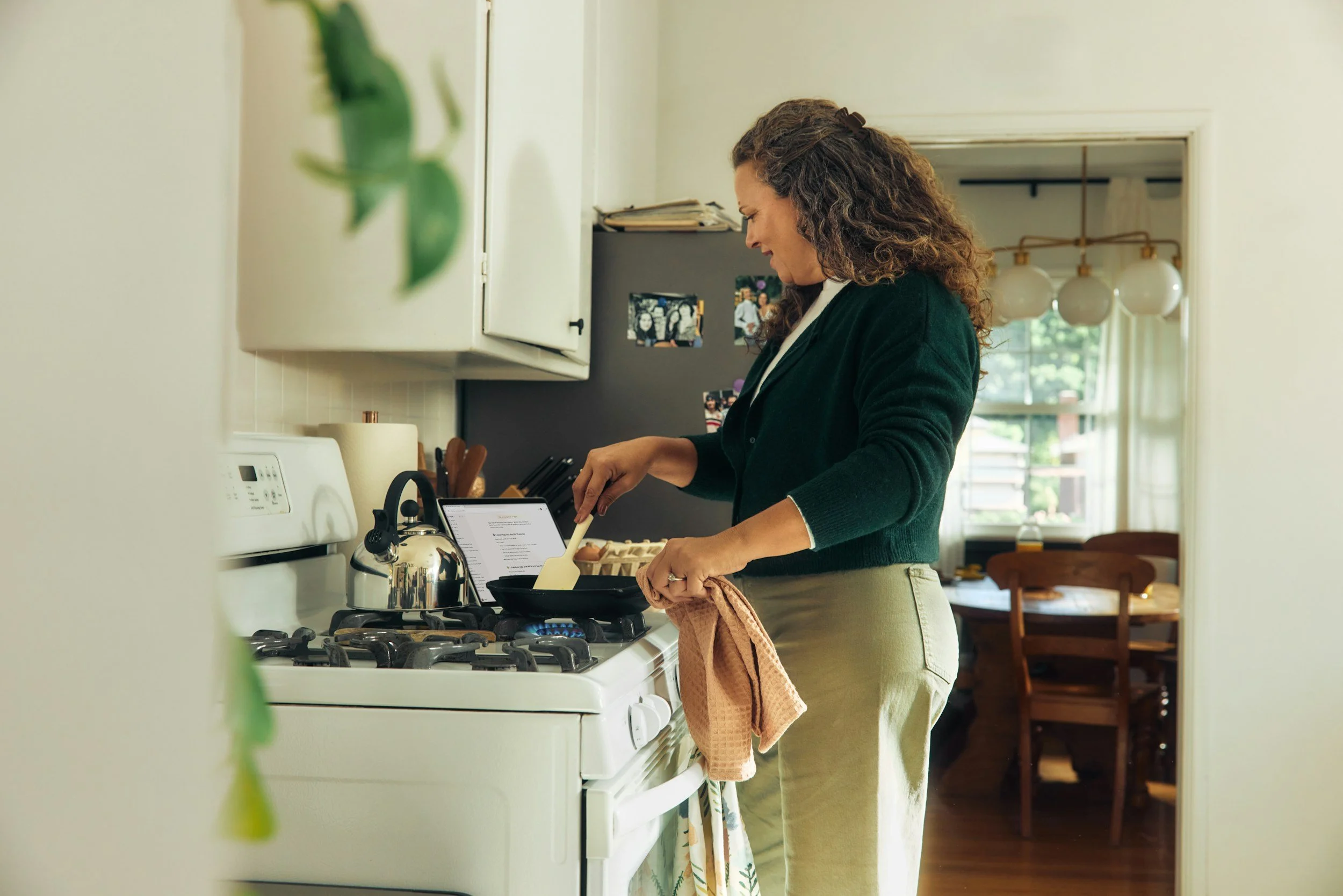 A woman with curly hair wearing a green cardigan and beige skirt cooking on a stovetop in a kitchen. She is smiling and looking down at food she is preparing. The photo is taken from a slightly side angle, with a fridge and dining area visible in the background.