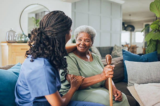 An elderly woman with gray hair smiling and holding a walking cane, talking to a caregiver with braided hair, in a cozy living room.