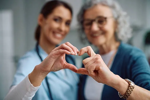 A young healthcare worker and an elderly woman making a heart shape with their hands, smiling at the camera.